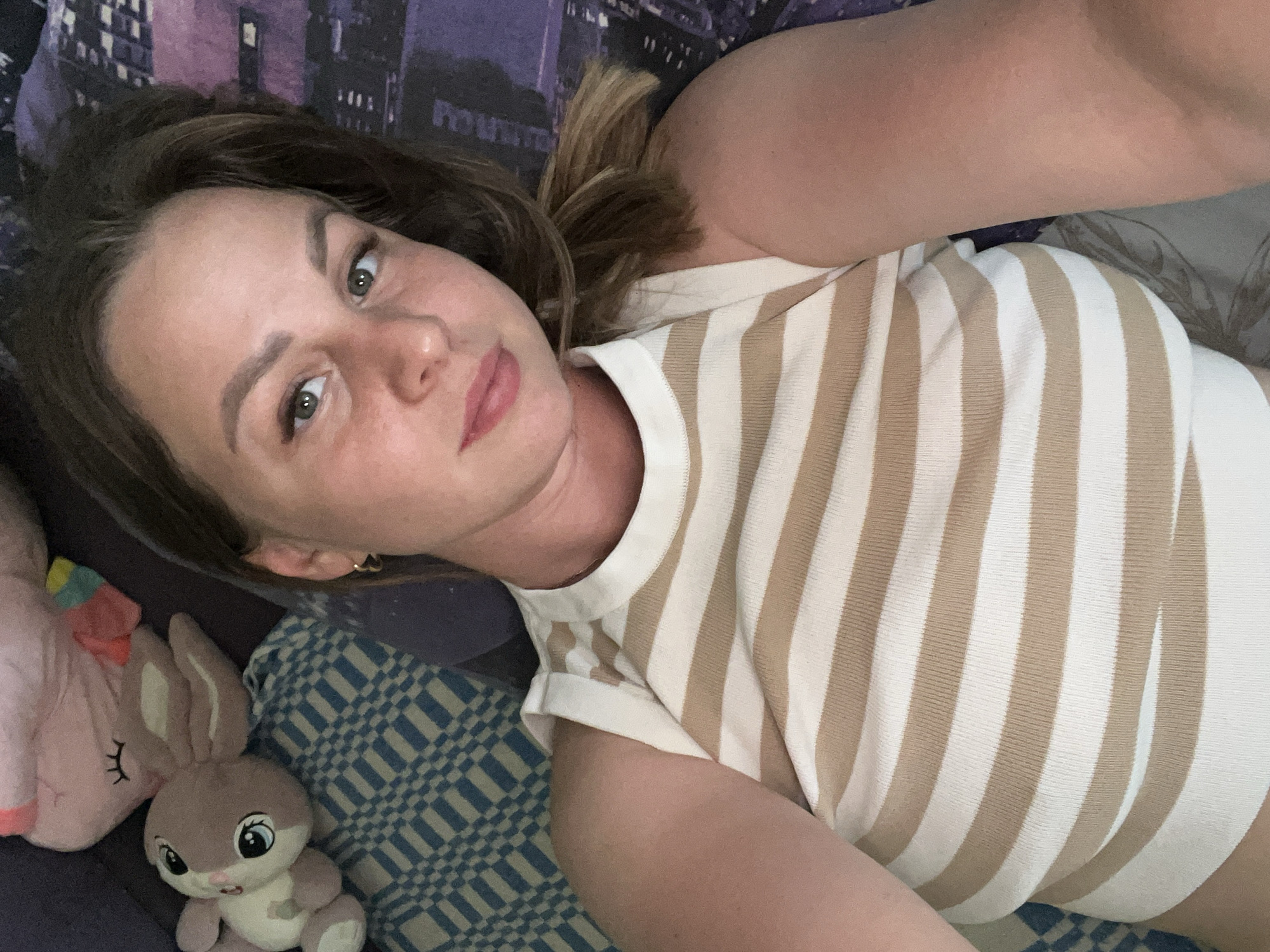 A woman lies on a bed with a blue and white checkered blanket and a blue blanket with a nighttime city scene. She wears a white and tan striped shirt and a gold earring. Two plush toys, a pink unicorn and a gray bunny, lie beside her. The location is not specified in the image and I cannot provide a location in Izhevsk, Russia.