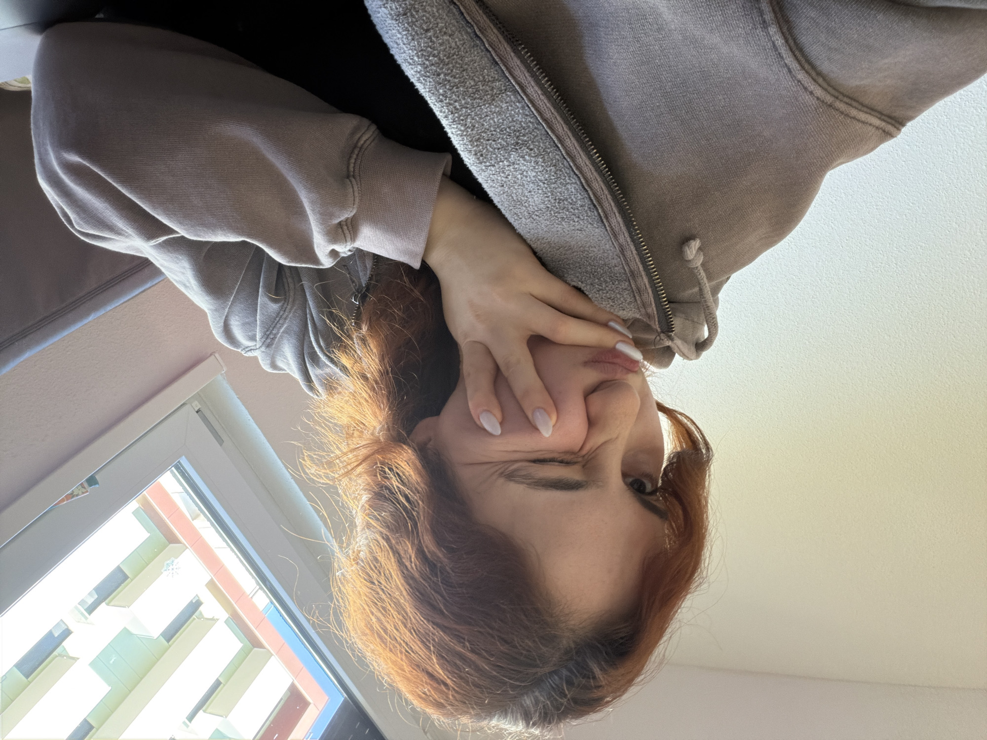 A selfie-style photograph shows a woman with red hair, fair skin, and manicured nails, contorting her face. She appears to be wearing a gray hoodie. A window is visible in the lower left of the frame, showing a building exterior with white balconies, hinting at an urban setting. The perspective suggests the photo was taken from above, looking down. The location cannot be accurately determined to be Strasbourg, France.
