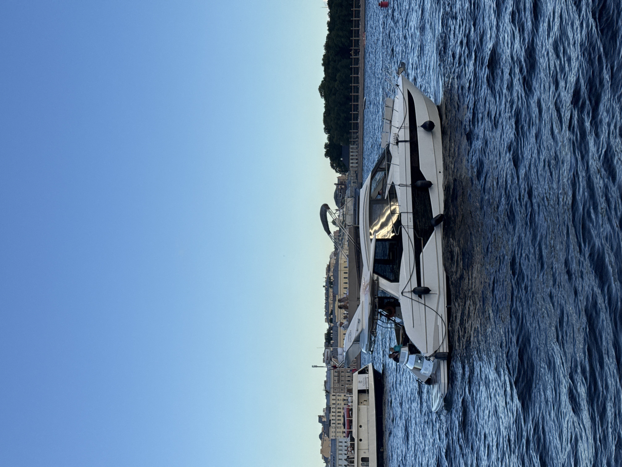A luxury white yacht is cruising on a river, likely the Neva River, in Saint Petersburg, Russia. The yacht has a sleek design with black trim and a hardtop roof. Several people are visible on deck, enjoying the ride. In the background, the historic cityscape of Saint Petersburg is visible, with its distinctive architectural style. The sky is a clear blue, indicating good weather. The water is a deep blue, with small waves. Another smaller boat is visible in the distance to the left of the yacht. The scene evokes a sense of leisure and opulence amidst the historical backdrop of the city.