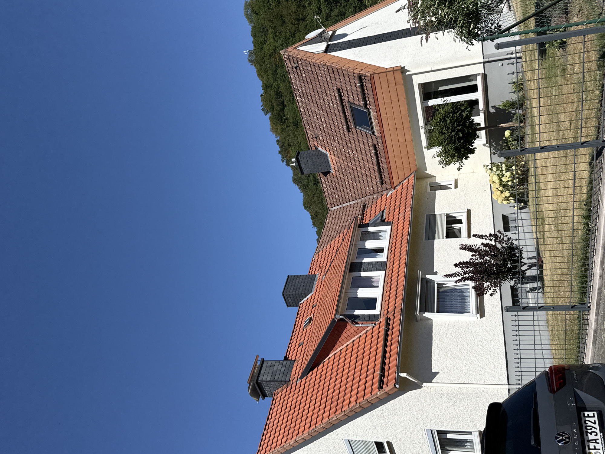 The image shows a two-story house with a red-tiled roof and white stucco walls under a clear blue sky. The house has multiple windows, some with roll-down shutters, and two dark grey chimneys. A portion of a dark grey Volkswagen Tiguan is visible in the foreground, parked on a paved surface. A metal fence with vertical bars separates the property from a dry, grassy yard where a small tree and bushes are planted. Behind the house, a lush green hillside is covered in trees, and a distant transmission tower is visible against the sky. The scene appears to be a residential area in the city of Bad Lauterberg im Harz, Germany, on a sunny day.