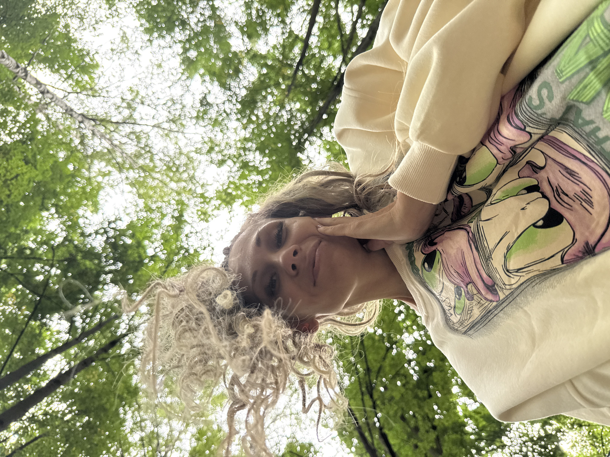 A low-angle, over-the-shoulder shot looks up at a young woman with curly blonde hair and light-colored clothing, her face partially obscured by her hand. She is outdoors, with a background of green trees and dappled sunlight. The image was captured in Rusanovo, Russia.