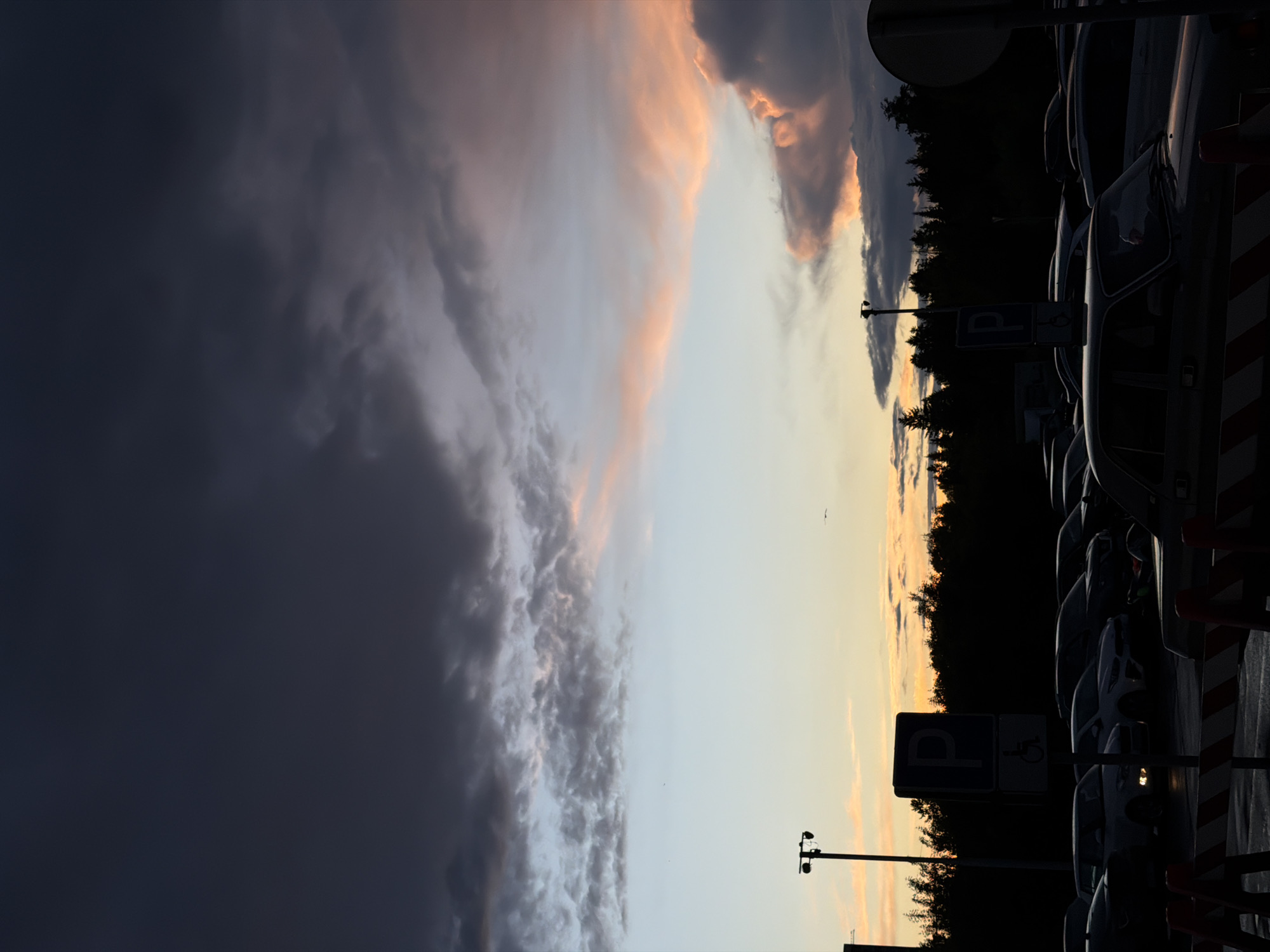 The image captures a scene in what appears to be a parking lot or parking garage. The location is implied to be Izhevsk, Russia. On the right side, there are rows of parked cars, mostly silhouetted. Parking signs are visible, including one with the international symbol of access. Towards the center, a line of trees separates the parking area from the sky. The sky displays a dramatic contrast with dark, stormy clouds on the left and a lighter, sunset-tinged area on the right. The lighting suggests either dusk or dawn. The overall mood is one of transition, with the weather and the light changing rapidly. No people are visible in the scene.