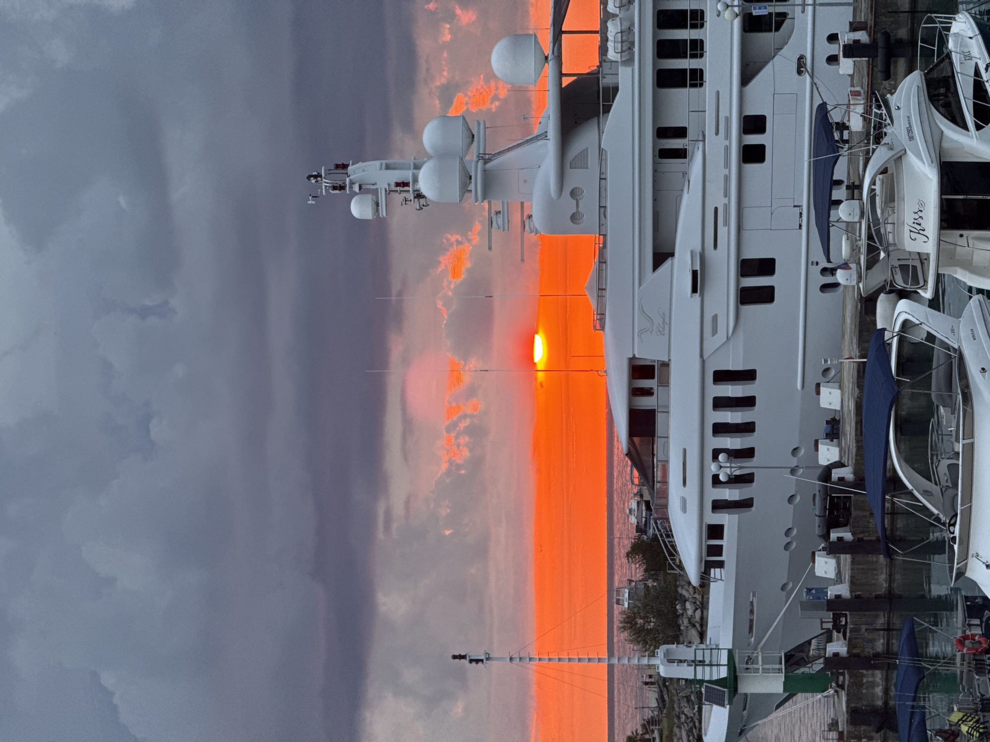 The image depicts a striking sunset over a harbor in Sochi, Russia. The sky is a dramatic mix of deep purples and vibrant oranges, with the setting sun casting a fiery glow. In the foreground, a large, white luxury yacht dominates the scene, its sleek lines contrasting with the textured clouds. Smaller boats are docked alongside it, some with blue canopies. The water is calm, reflecting the colors of the sky. The scene appears to be at dusk, with artificial lights from the boats and harbor infrastructure beginning to emerge. The overall impression is one of opulence and natural beauty.