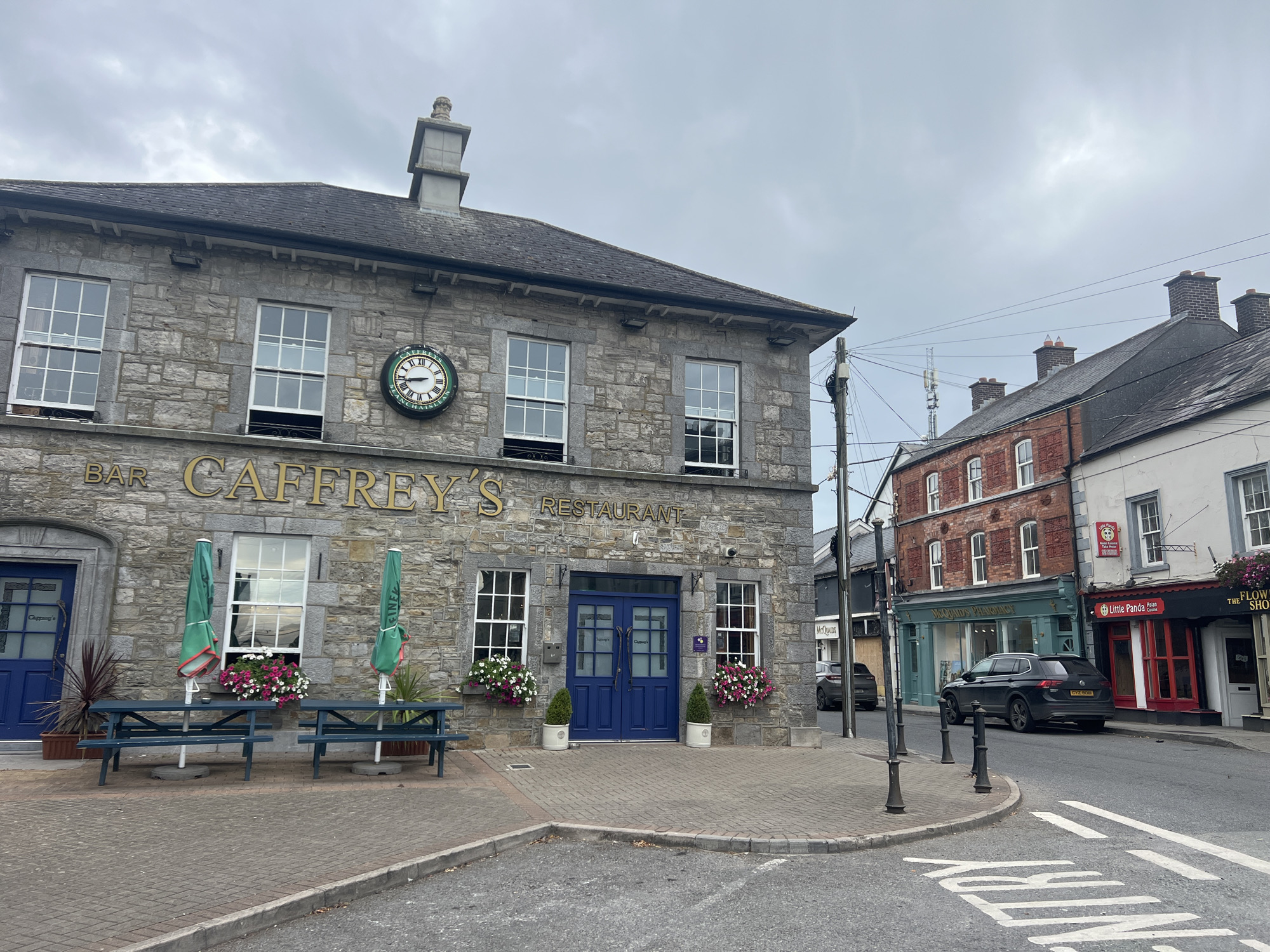 A building with stone exterior features a sign that reads "BAR CAFFREY'S RESTAURANT". Two green umbrellas stand over blue picnic tables on a paved area in front of the building. Two large blue double doors are centrally located on the building's facade, flanked by windows. A clock with Roman numerals is mounted on the upper part of the building. Across the street, a row of brick buildings houses various businesses, including a pharmacy and a restaurant with red and black signage. A dark-colored SUV is parked on the street, and a white car is visible further down. The sky is overcast.