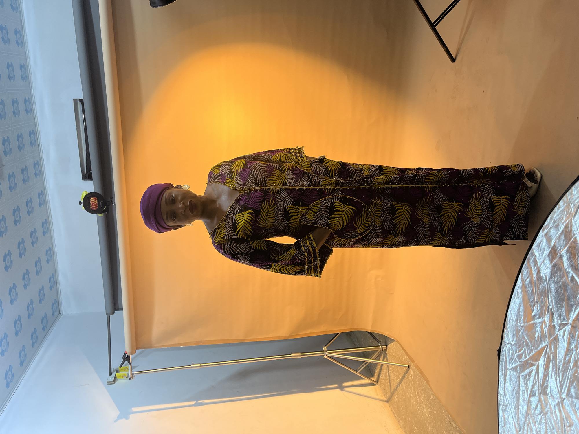 A woman wearing a purple head covering and a patterned long dress with green and purple leaf designs is posing for a photo in a studio setting. She is standing in front of a backdrop with an orange gradient. Studio lighting equipment, including reflectors and stands, are visible in the scene. The walls of the studio are decorated with a blue floral pattern. The photo was taken in the city of Gura Top, Nigeria.