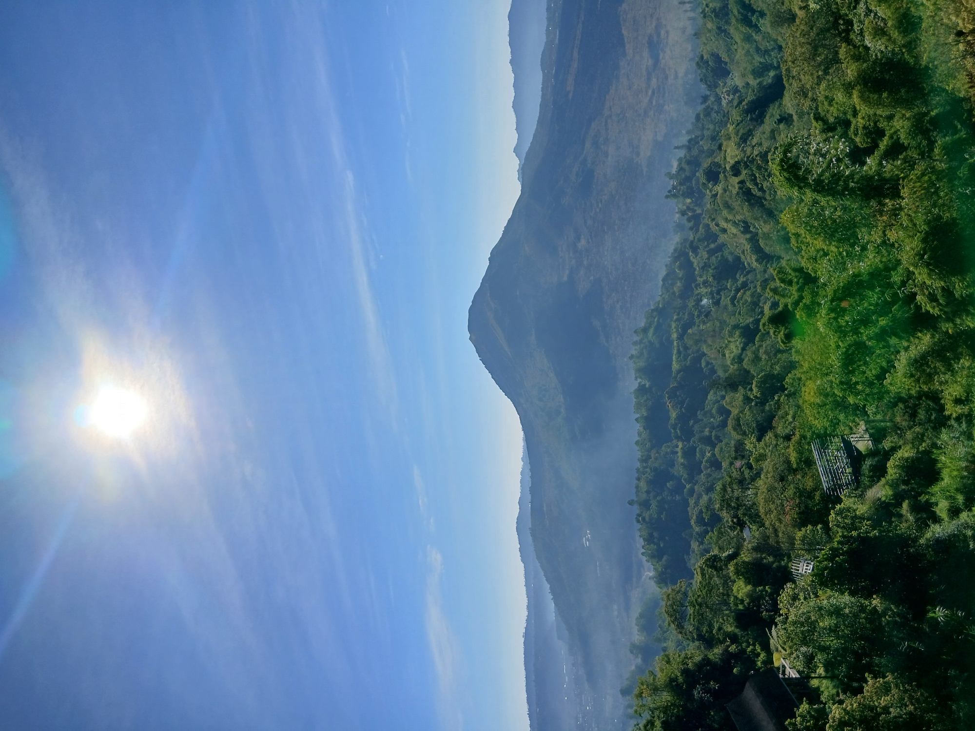 This vertical, eye-level, medium-wide shot captures a serene landscape at the city of Kintamani, Indonesia. The sky is a vibrant blue, with the sun shining brightly near the top left, casting a slight lens flare and light rays across the frame. Wispy clouds stretch across the upper portion of the sky.
In the mid-ground, a large, conical mountain dominates the horizon, its slopes shrouded in a light mist. To the left of this prominent mountain, smaller, rolling hills recede into the distance, also softened by haze. The middle section of the image is densely packed with lush green trees, creating a textured, undulating terrain. A few structures, including what appears to be a building with a dark roof and some metal scaffolding or structures, are visible nestled within the foliage.
The foreground is also filled with dense greenery, with various shades of green indicating different types of trees and vegetation. The overall impression is one of natural beauty and tranquility, with the mist adding a sense of depth and atmosphere to the mountainous terrain. There are no people or animals visible in the frame.