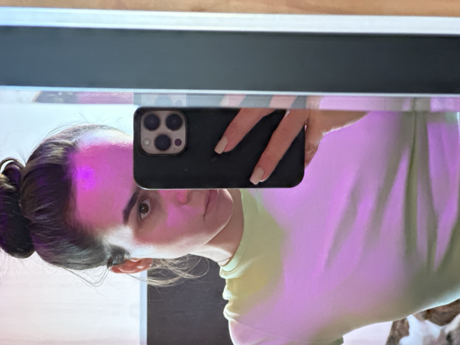 A female subject is depicted in a mirror selfie taken in an indoor setting within the city of 3-Y Uchastok, Russia. The individual, an adult with brown hair tied in a bun, has visible brown eyes and wears a light green short-sleeved top. Her right hand, with light-colored manicured fingernails, holds a dark-colored smartphone with a triple camera array, positioning it to capture her reflection. A distinct pink-purple light source illuminates the subject's forehead and upper torso. The environment is minimally visible, showing a dark vertical frame or object reflected on the right edge of the mirror, and a lighter, possibly wooden surface further to the right. A darker, undefined background element is reflected behind the subject's left shoulder. The subject's gaze is directed towards the smartphone camera.