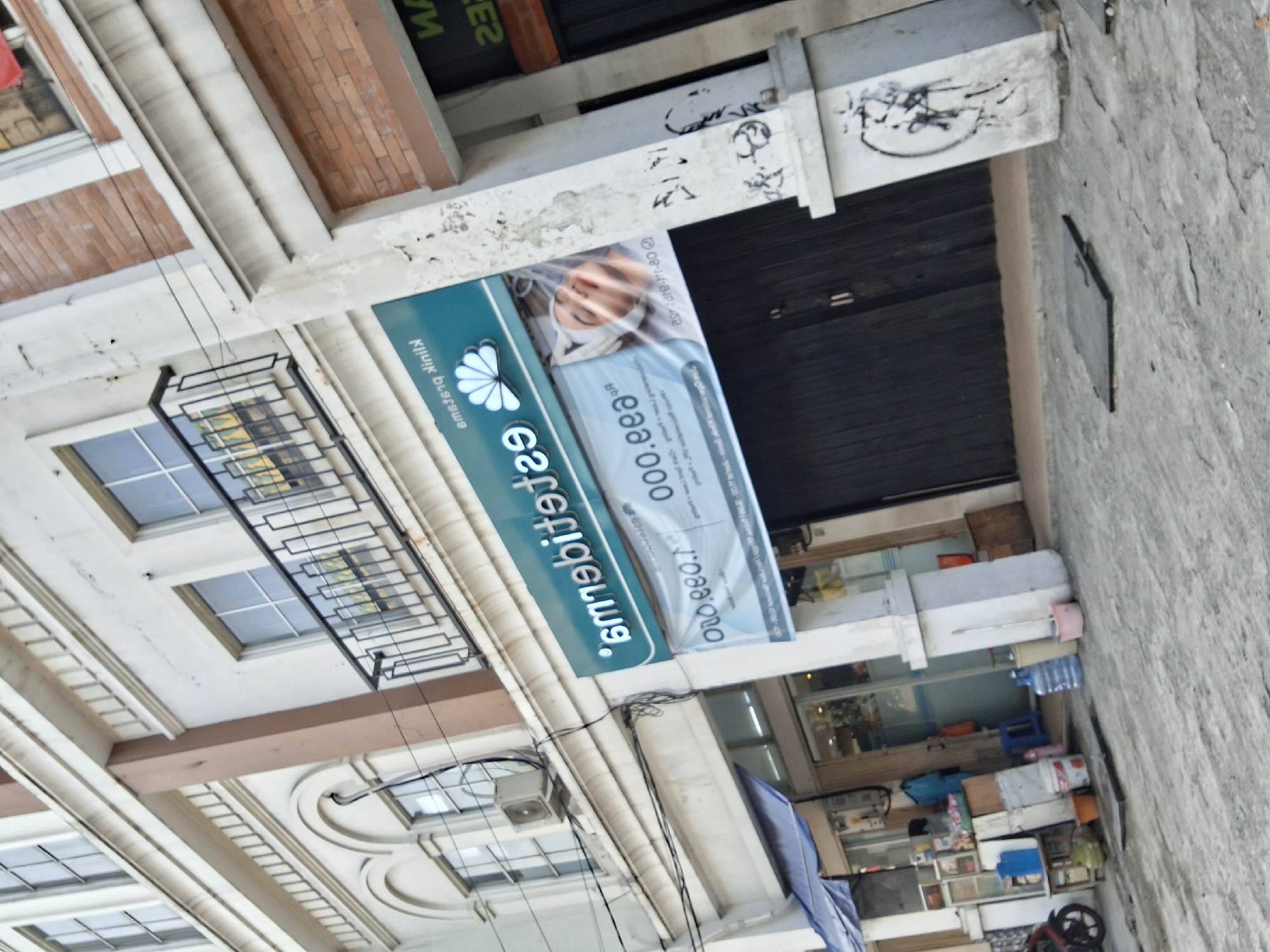 The image depicts the exterior of a commercial building with multiple storefronts in South Jakarta, Indonesia. The building features a white facade with arched windows and decorative molding on the upper levels. A prominent teal-colored sign reads "estheters" in white lettering, with a stylized fan logo. Below this sign, a banner displays an image of a person's face with a white head covering, accompanied by text in Indonesian and possibly English, including what appears to be a phone number.
On the ground floor, there are several dark-colored storefronts. To the left, a blue awning covers part of a storefront. Various items are visible outside this storefront, including a motorcycle, several buckets, plastic containers, and a green spherical object, possibly a coconut. Further to the right, a large white pillar stands on the sidewalk, adorned with black graffiti. In front of a storefront to the right of the pillar, a dark rectangular metal cover is partially visible on the ground. The street or sidewalk area is paved and appears somewhat worn. The overall scene suggests a commercial area with typical street-level activity.