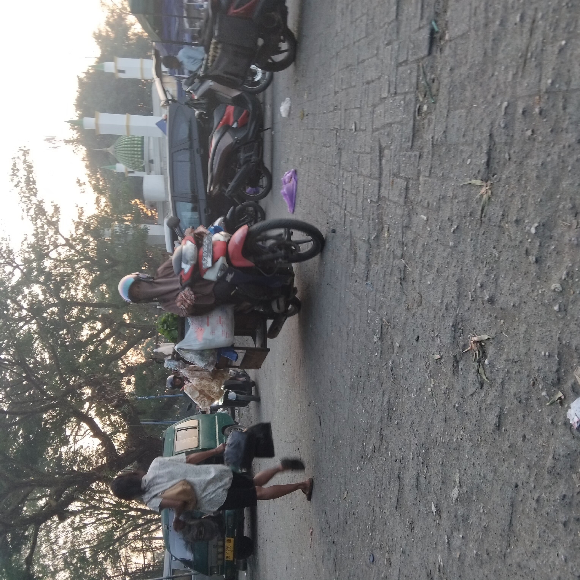 In Banjarbaru, Indonesia, a street scene unfolds with several individuals and vehicles. A woman wearing a brown hijab and a beige outfit rides a red and white scooter, carrying a large white plastic bag filled with goods and a smaller brown bag. She is positioned in the center of the frame, on a paved surface. To her left, a person wearing a light-colored short-sleeved shirt and dark shorts walks away from the camera, carrying two dark bags and a light-colored shoulder bag. They are walking next to a green van with license plate DA 1205 BB. Further to the left, a portion of a dark-colored SUV is visible. In the background, a mosque with green domes and white minarets stands against a hazy sky. Trees are scattered throughout the scene, and other scooters and market stalls are partially visible. The ground is a mix of rough pavement and dirt.