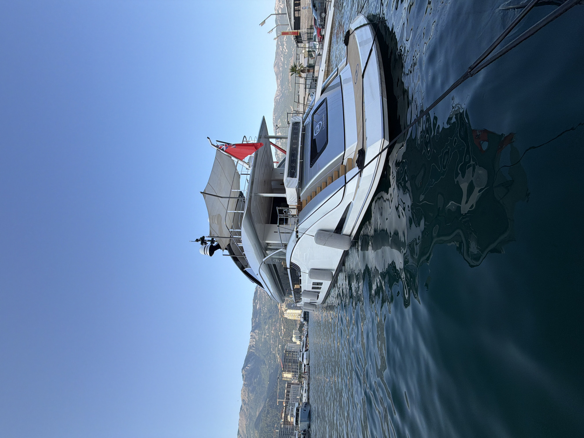 The image shows a luxurious white superyacht docked in a marina. The yacht features multiple levels, a red flag on top, and several grey fenders along its side. The stern of the yacht is visible, with the name "JETSETTER" and "LONDON" printed on it. The water is a deep blue-green, and its surface reflects the yacht and the sky. In the background, a city skyline with buildings and mountains can be seen under a clear blue sky. The marina appears to be located in Budva, Montenegro, given the distinctive mountainous backdrop often associated with the region. There are no people visible in the image. The scene depicts a serene and opulent maritime setting.