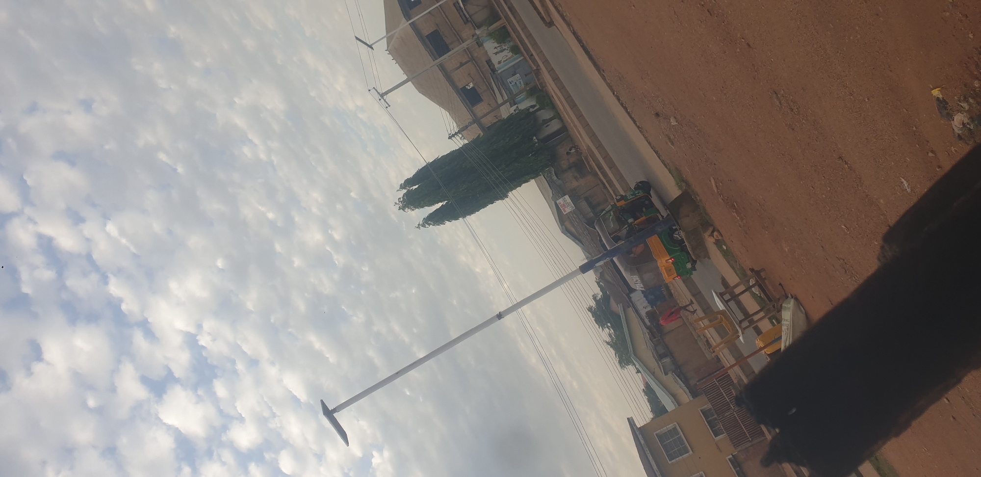 The image depicts a street scene in Jos, Nigeria, under a cloudy sky. A tall, white street light with a solar panel is prominently featured on the left. In the mid-ground, a green and yellow motorized tricycle, commonly known as a "keke," is driving on the paved road. A person in a red shirt is walking on the side of the road. Several buildings line the street, some appearing to be residential with windows visible. Tall, slender trees stand to the right of the street light, adding to the urban landscape. The foreground is dominated by reddish-brown dirt and debris. The overall atmosphere suggests a typical day in a Nigerian city, with a blend of infrastructure, transportation, and everyday activities.