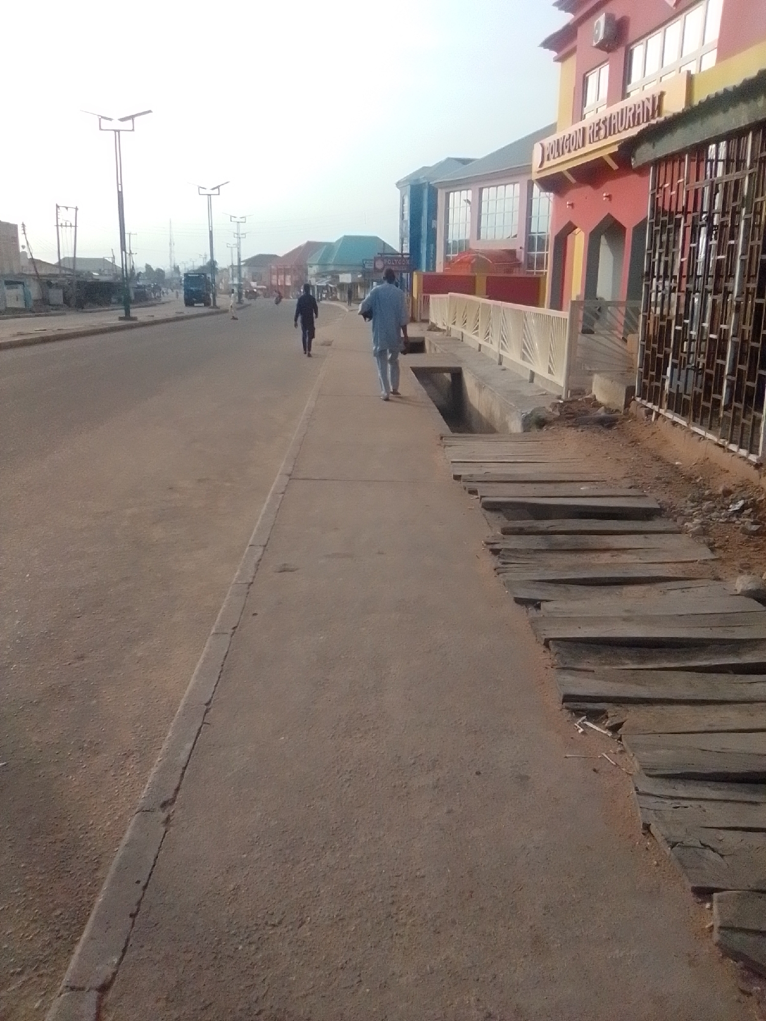 The image shows a street scene in Pantame, Nigeria, with a focus on a sidewalk and the road. On the sidewalk, two individuals are walking away from the camera. The person on the left is wearing a dark garment, while the person on the right is wearing a light blue garment. To the right of the sidewalk, there is a building with a sign that reads "POLYGON RESTAURANT". The building has a red and yellow facade and multiple windows. Adjacent to the restaurant is a metallic grate structure. The sidewalk itself is concrete, and next to it, a series of wooden planks are laid out, some appearing worn and uneven. The road in front of the sidewalk is paved and appears to be a busy thoroughfare, with streetlights visible in the distance. The sky is overcast, suggesting a hazy or possibly early morning/late afternoon light.