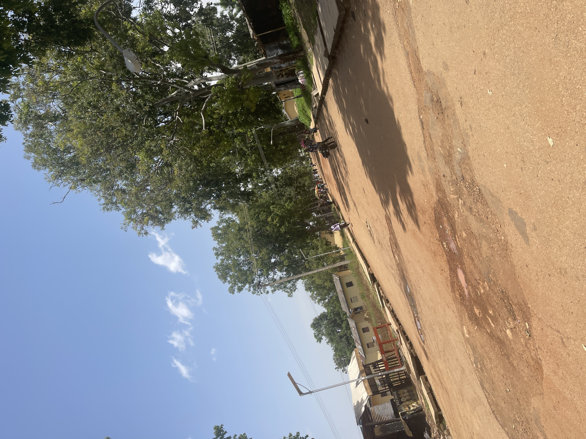 The image depicts a street scene in Rafin Par, Nigeria, under a clear blue sky with scattered clouds. The unpaved road, composed of reddish-brown dirt and gravel, shows signs of wear and tear, with visible puddles and patches of mud. On the left side of the road, several low-rise buildings with corrugated metal and tiled roofs are present, some appearing weathered and with sparse vegetation nearby. A prominent street lamp with a modern solar panel fixture stands on the left.
The right side of the road is lined with mature trees that cast shadows across the path. A paved sidewalk runs alongside these trees, bordered by concrete blocks. Further down the street, several people are visible. Two individuals are riding on a single motorcycle, while another person is walking. In the distance, more people and motorcycles are present, suggesting ongoing activity on the street. The overall environment appears to be a residential or mixed-use area within the city, characterized by its unpaved road and vegetation.