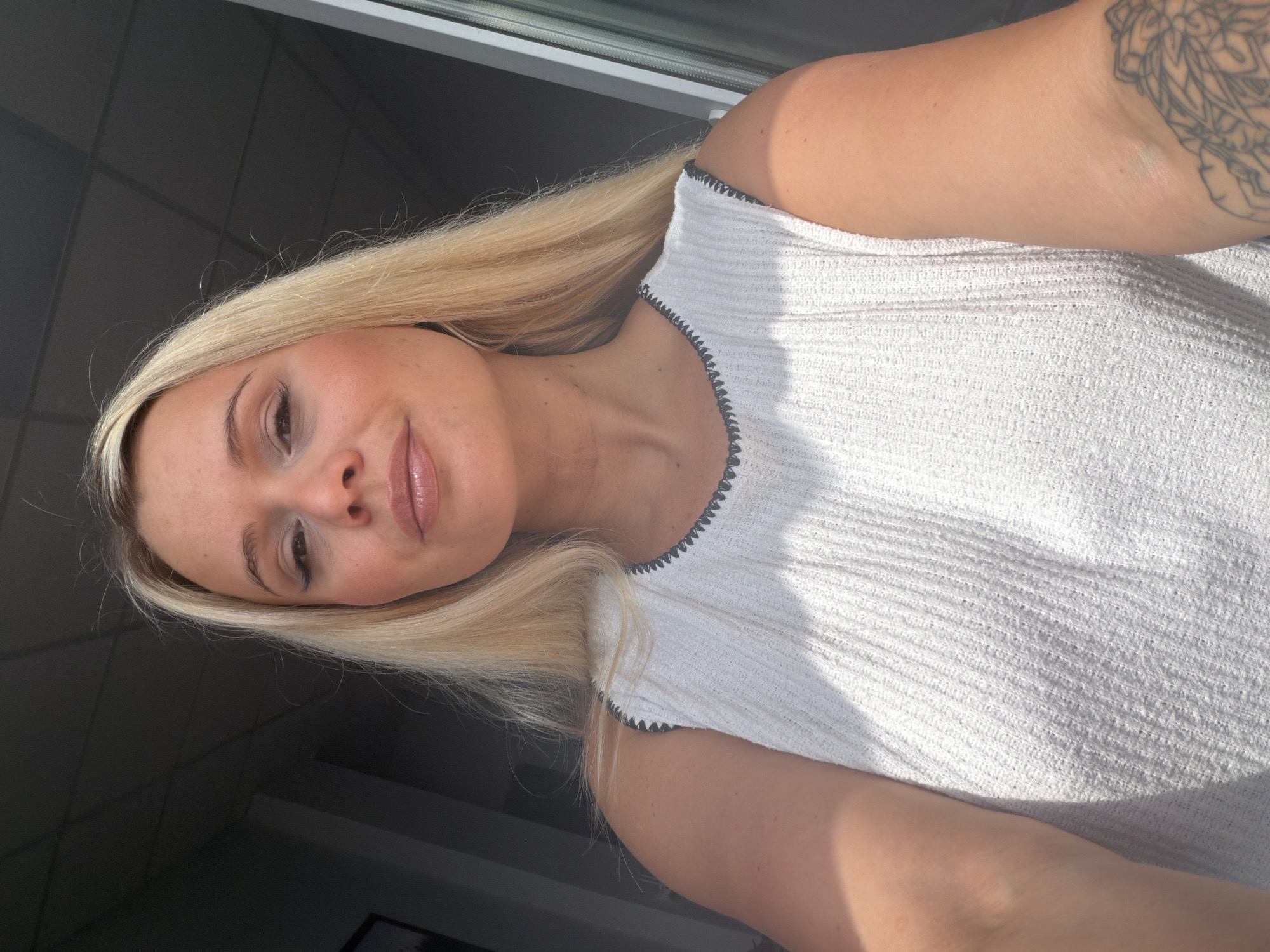 A blonde woman with fair skin and makeup is shown in a close-up selfie, looking towards the camera. She has light brown lipstick and eyeliner. She is wearing a white sleeveless top with a textured pattern and a dark trim around the neckline. Her left arm, visible on the right side of the image, has a tattoo of a mandala. The background appears to be an indoor setting with dark walls and a window with vertical blinds. The lighting suggests it is daytime. No specific landmarks or elements within the image can be used to definitively place this scene in Sarapul, Russia.