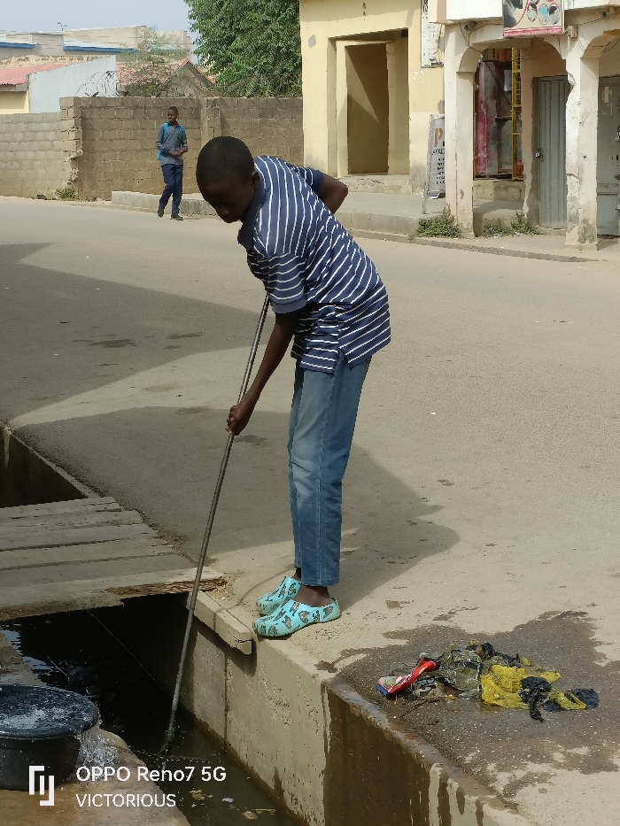 The image displays an outdoor street scene in Dutse, Nigeria, under clear daylight. A young male, wearing a blue and white striped short-sleeved polo shirt, light blue jeans, and light blue patterned clogs, is positioned in the foreground, bent forward over a concrete drainage ditch. He uses a long metal rod, held with both hands, to clear or stir debris within the water-filled ditch. Adjacent to him, on the paved ground beside the ditch, rests a collection of wet refuse, including yellow and black plastic bags and a red cylindrical object, possibly a can. A dark bucket, seemingly overflowing into the ditch, is partially visible in the lower left. Further down the ditch, multiple wooden planks span across it. In the middle ground, another male, dressed in a grey long-sleeved shirt and dark trousers with a backpack, walks away from the viewer on the paved road. The background is composed of light-colored, single-story buildings with various doorways and architectural details, some suggesting commercial use, and a low brick wall topped with barbed wire. Sparse green vegetation is visible behind the structures.