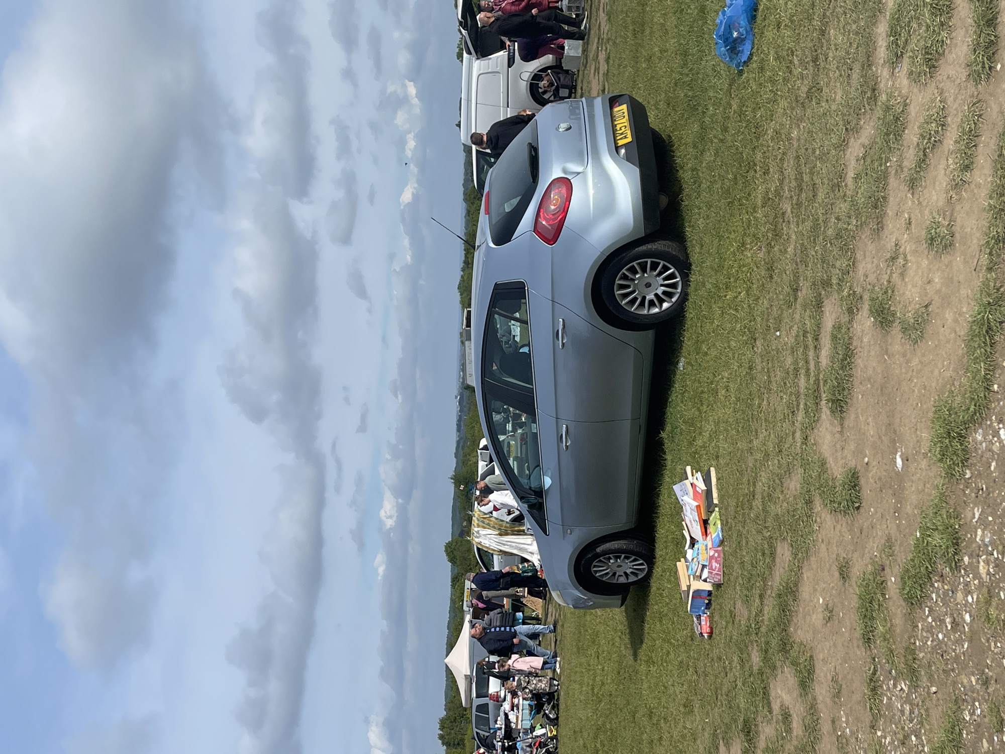A photograph shows a silver Fiat Bravo parked on grass next to a collection of books, likely part of a car boot sale or similar event. Other vehicles, including a white van, and various stalls or pitches can be seen in the background. Several people are present, suggesting a market or fair atmosphere. The photograph seems to be taken outdoors, possibly in a field or open space. The UK license plate "AK57 ODV" can be clearly seen on the car. Based on the search results, this image was captured in Sevenoaks, United Kingdom.