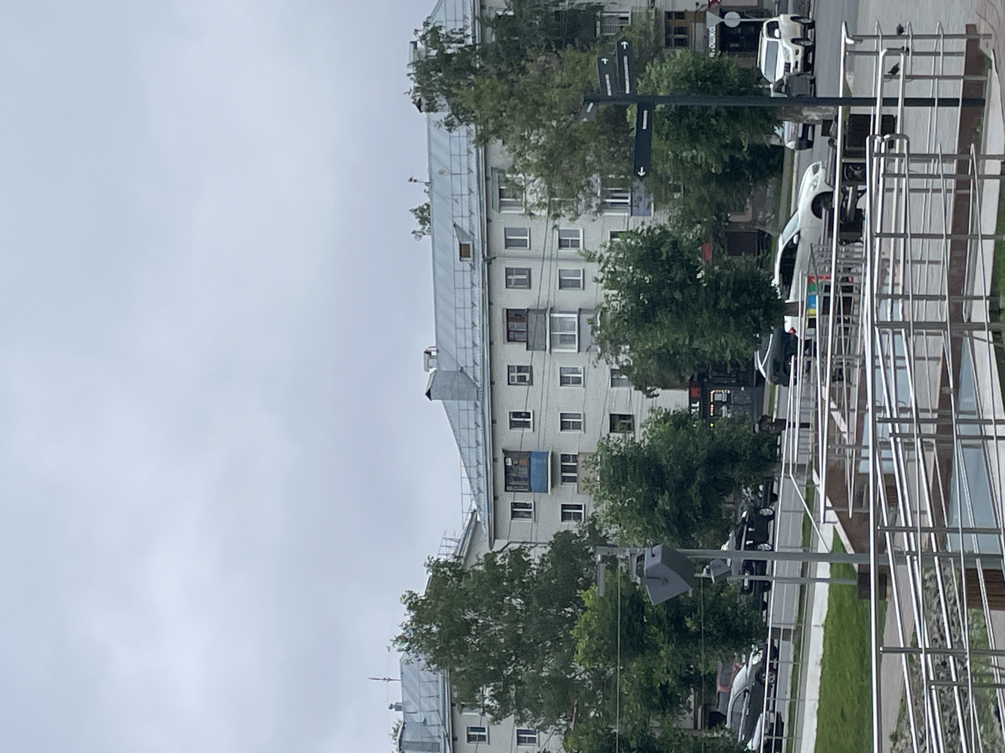 The image shows a view of a street in Kirov, Russia. In the foreground, there are metal railings and a ramp leading down. Behind the railings, a grassy area is visible, followed by a street with several cars driving and parked. On the other side of the street, there are several multi-story apartment buildings with gray roofs and light-colored walls. Trees are interspersed between the buildings and along the street. A traffic signpost with directional arrows is also visible. The sky is overcast and gray.