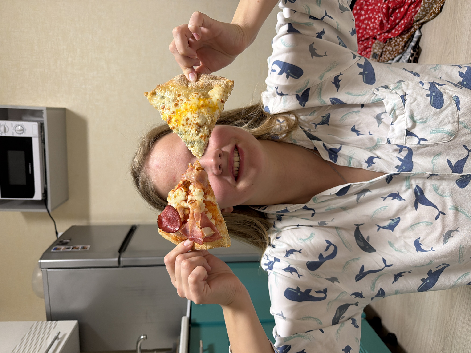 A woman is holding two slices of pizza up to her face, one over each eye. She is wearing a white shirt with blue whale patterns. The background shows a kitchen setting with a microwave, a silver refrigerator, and a sink. The environment appears to be indoors. The location is Izhevsk, Russia.