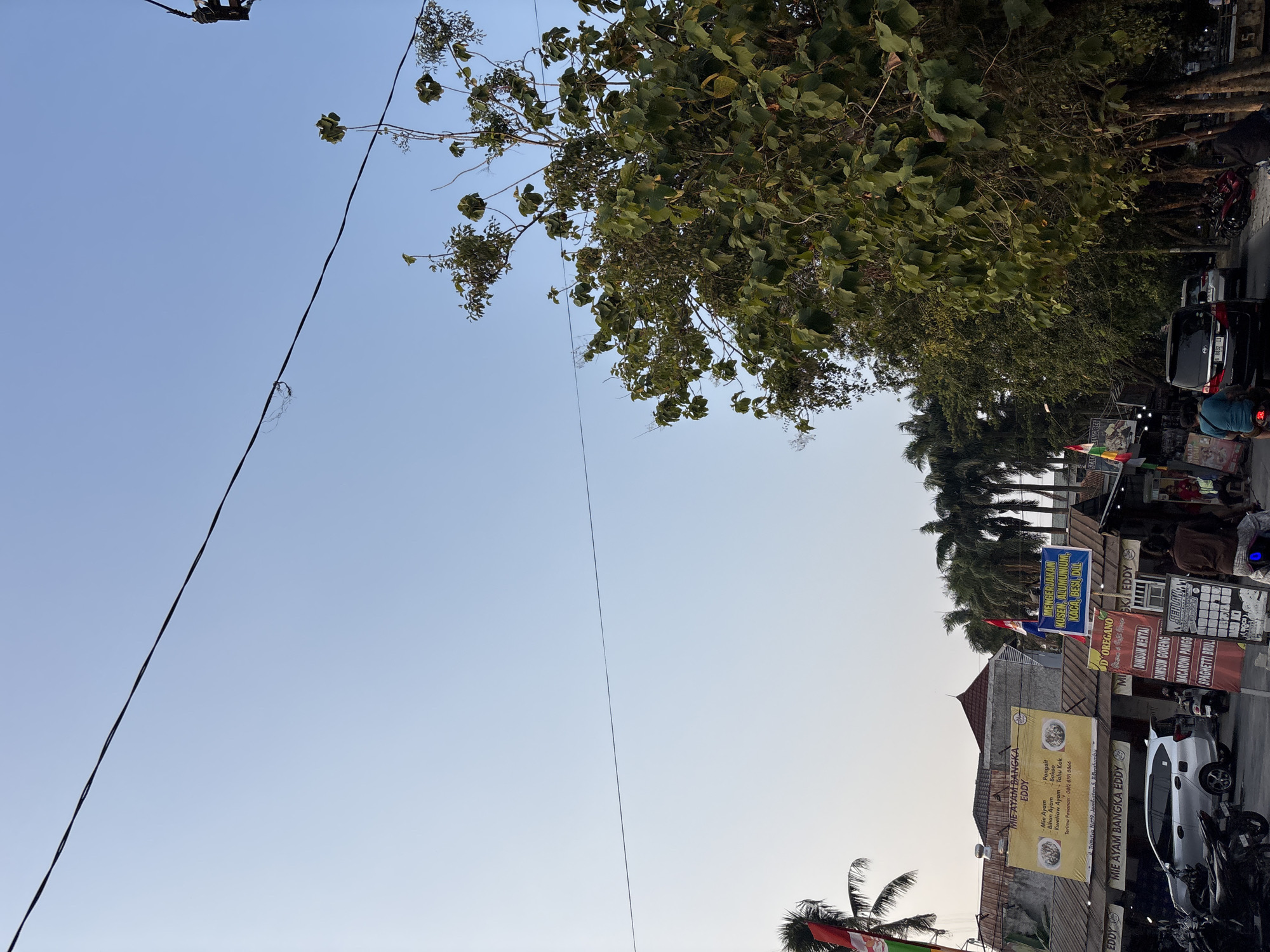 The image depicts a street scene in Bekasi, Indonesia, characterized by a clear blue sky with power lines traversing across it. On the left side, several buildings are visible, featuring yellow and blue signs advertising food establishments, specifically "MIE AYAM BANGKA EDDY." Palm trees and other foliage line the street, with a large tree dominating the right portion of the frame. Vehicles, including cars and motorcycles, are parked and in motion. People are present, some riding motorcycles and others appearing to be pedestrians. The overall impression is that of a bustling commercial street in a tropical urban environment.