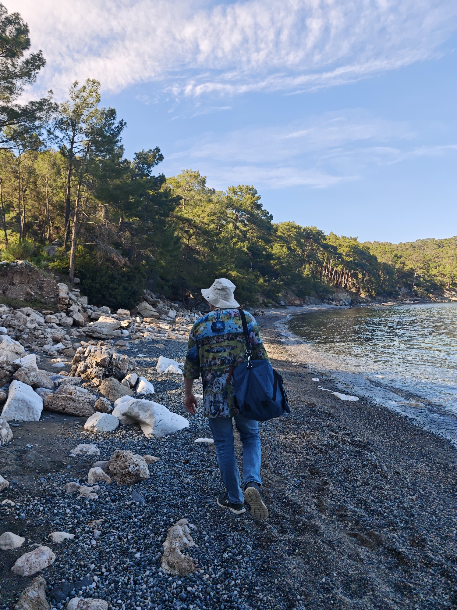 An individual is captured from behind, walking along a rocky and pebbly beach next to a calm sea under a bright sky. The scene is set in Kemer, Turkey, featuring a distinctive Mediterranean coastline.
The person, appearing to be an adult, wears a light-colored bucket hat, a multi-colored patterned shirt with shades of blue, green, and brown, blue jeans, and dark sneakers with white soles. They are carrying a dark blue duffel-style bag over their left shoulder. Their posture suggests a leisurely walk, with one foot visible mid-stride on the pebbled ground.
To the left of the person, the beach is covered with a mix of large, irregularly shaped white and light brown rocks, and smaller dark pebbles. Further inland, a dense forest of tall, green pine trees covers the rising terrain, their foliage catching the sunlight. The right side of the image shows the tranquil, clear waters of the sea, with gentle ripples near the shore. The seabed appears dark through the shallow water.
The sky is a vibrant light blue with scattered white, wispy clouds, suggesting fair weather. The quality of light, with warm tones on the trees and rocks and some visible shadows, indicates either early morning or late afternoon, often referred to as the golden hour. No specific text is visible in the image. The overall impression is one of peaceful exploration in a natural coastal environment.