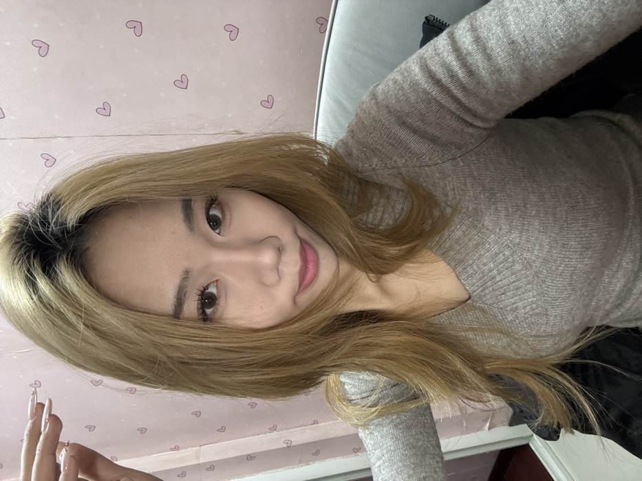 A young woman with tan skin and long, blonde hair looks toward the camera. She is wearing a gray, long-sleeved shirt with a V-neck, and her long fingernails are visible. The background features a light pink wall adorned with small heart-shaped designs, while a partially visible light-colored sofa is behind her.