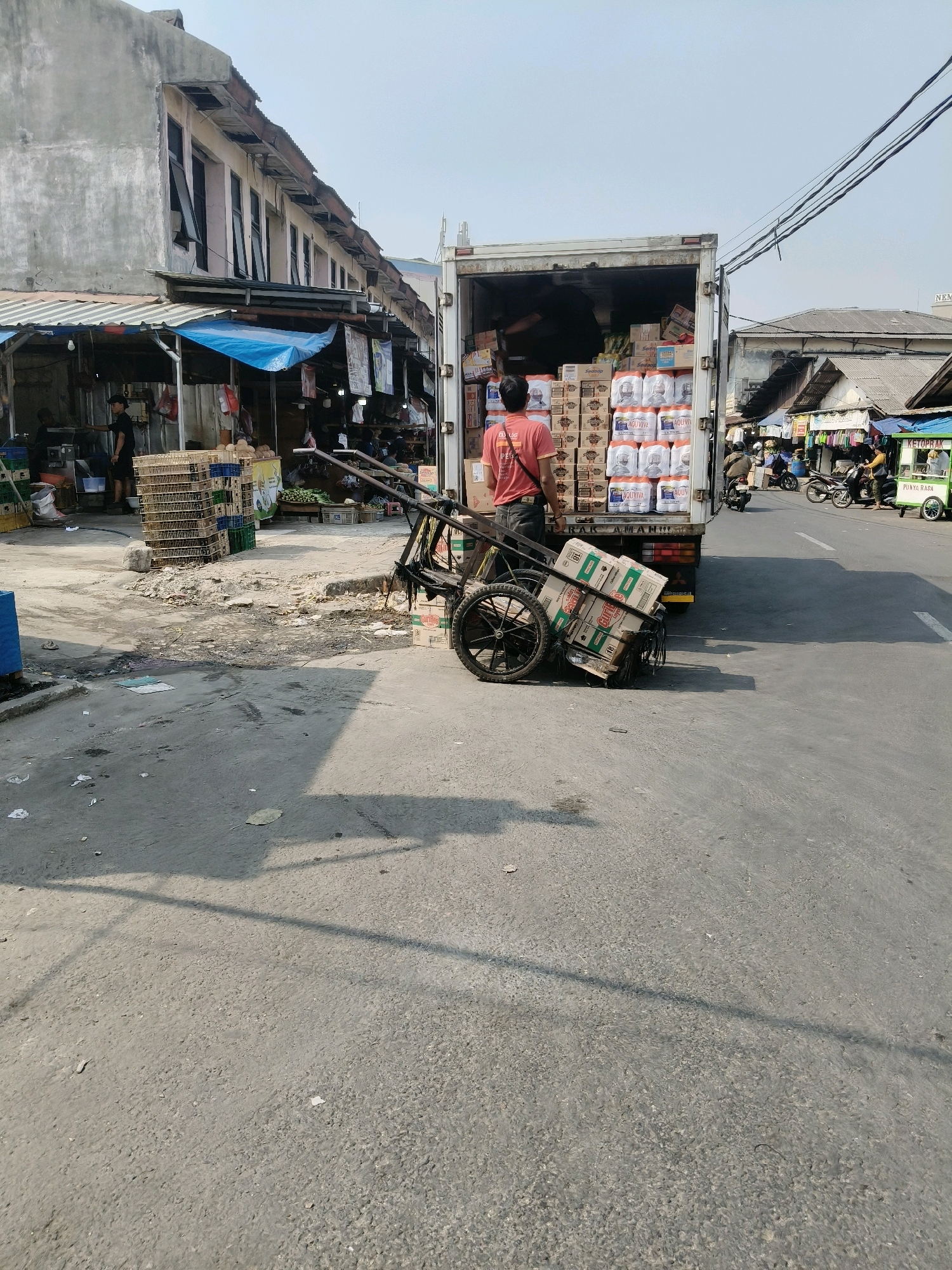 A man in a red shirt and black pants is standing next to a truck loaded with boxes and a two-wheeled cart also laden with boxes. The truck is parked on an asphalt road in a market area. Buildings with shops and stalls line the left side of the road, and more buildings and a small green food cart are visible on the right. The sky is clear and blue. The scene is likely in South Tangerang, Indonesia, judging by the context of the market environment typical of the area.