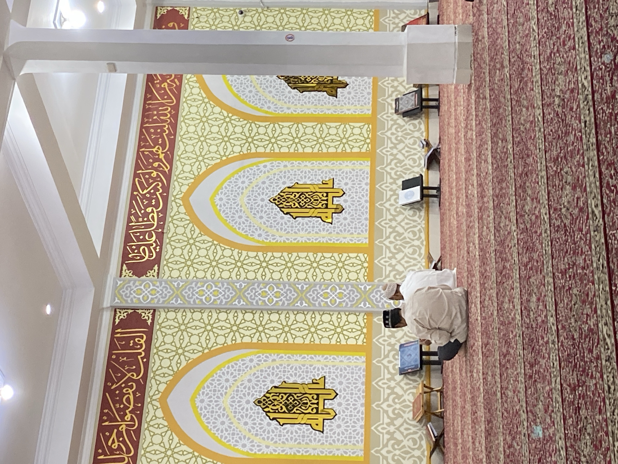 The image depicts the interior of a mosque in Lhokseumawe, Indonesia, featuring two men seated on a red carpet. The men are facing away from the camera, engaged in prayer or reading. The mosque's walls are adorned with intricate Islamic calligraphy and geometric patterns in shades of gold, maroon, and cream. Arched windows with decorative grilles are visible on the wall behind the men. Several religious books are placed on stands around the prayer area. The overall ambiance suggests a serene and spiritual setting within the mosque.