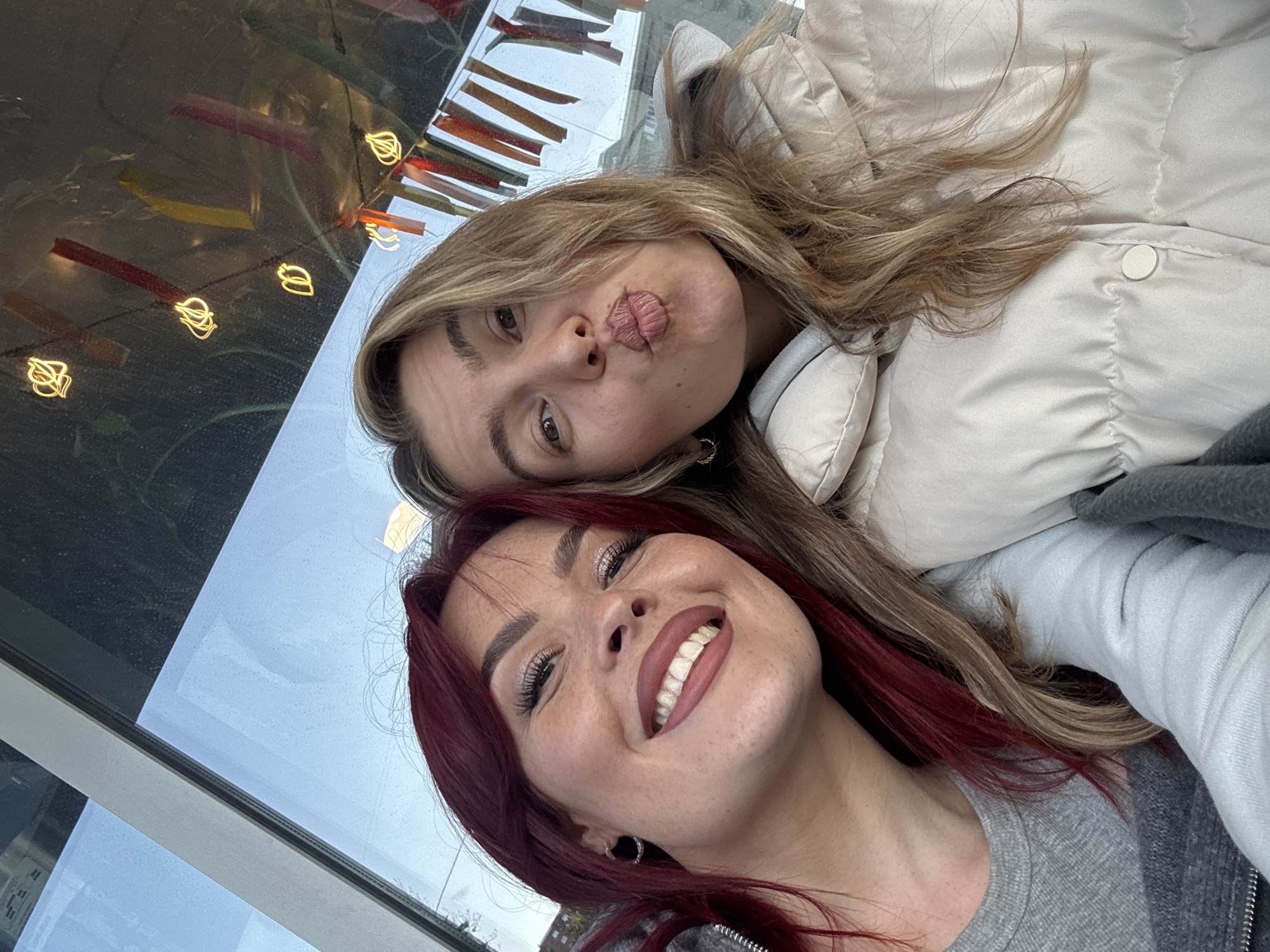 A tilted, close-up selfie features two young women. The woman in the foreground on the right has long, rich red hair and a broad, joyful smile, revealing her upper teeth. She wears shimmering eye makeup with long eyelashes and reddish lip liner. Her attire includes a grey ribbed top and a darker grey zippered jacket, with a silver hoop earring visible.
Behind her, to the left, is another woman with long blonde hair. She poses playfully with puckered lips, looking towards the camera, while wearing a light beige puffer jacket.
The background suggests an indoor setting next to a large window. Through the window, a light blue sky is visible, indicating daytime. The window also reflects several small, golden, stylized leaf-shaped lights, which appear to be indoor decorations. The overall mood is cheerful and friendly. No specific location cues or text are visible in the image.