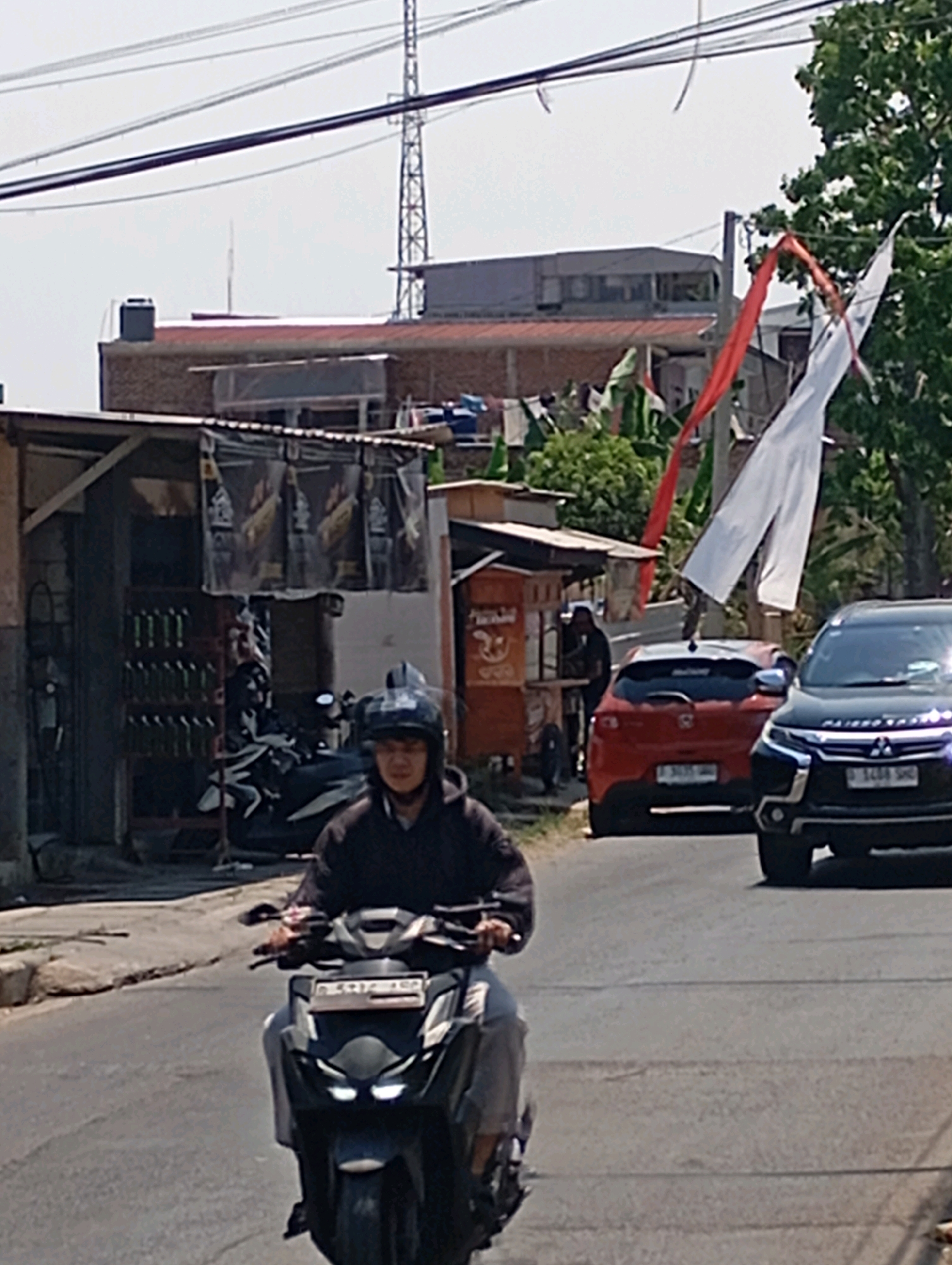 In Cimahi, Indonesia, a person wearing a dark hoodie and helmet rides a dark-colored scooter on a paved road. Behind the scooter, a red hatchback and a dark SUV are visible. To the left of the road, there is a small structure with merchandise displayed on shelves, possibly a roadside shop. In the background, buildings and trees line the street, with power lines and a tall communication tower overhead. Red and white flags are seen fluttering in the wind, strung between poles and trees. The sky is clear and bright, suggesting a sunny day.