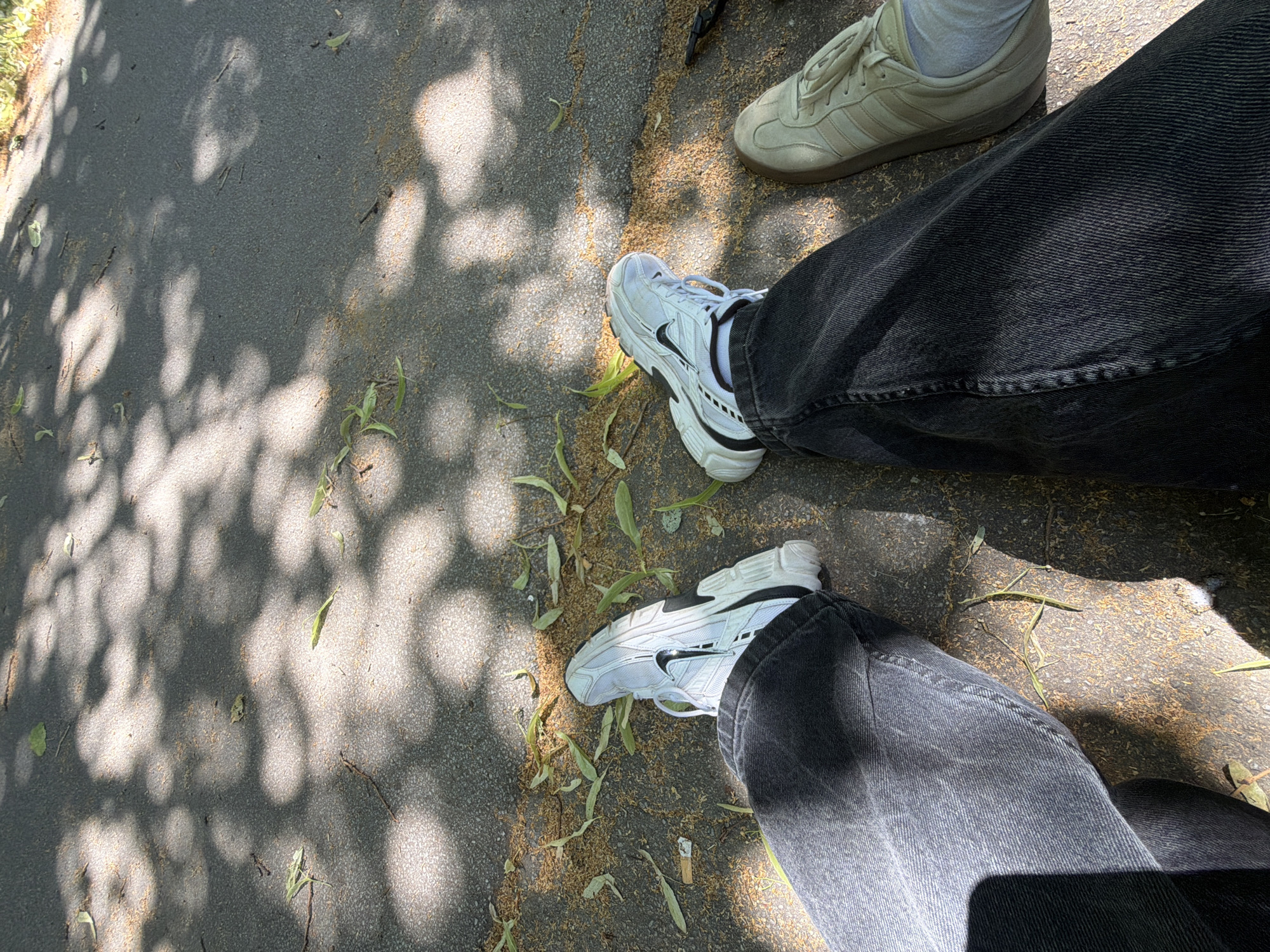 The image shows a close-up view of two pairs of legs and feet, both wearing casual footwear and dark denim pants, resting on a paved surface outdoors. The pavement is covered with scattered leaves and debris, and dappled sunlight creates a pattern of light and shadow across the ground. One person is wearing white Nike sneakers with black accents, while the other is wearing light beige sneakers. The scene suggests a relaxed outdoor setting, possibly a park or a street in a city. There are no discernible landmarks or contextual clues that would specifically place this scene in Dorsten, Germany.