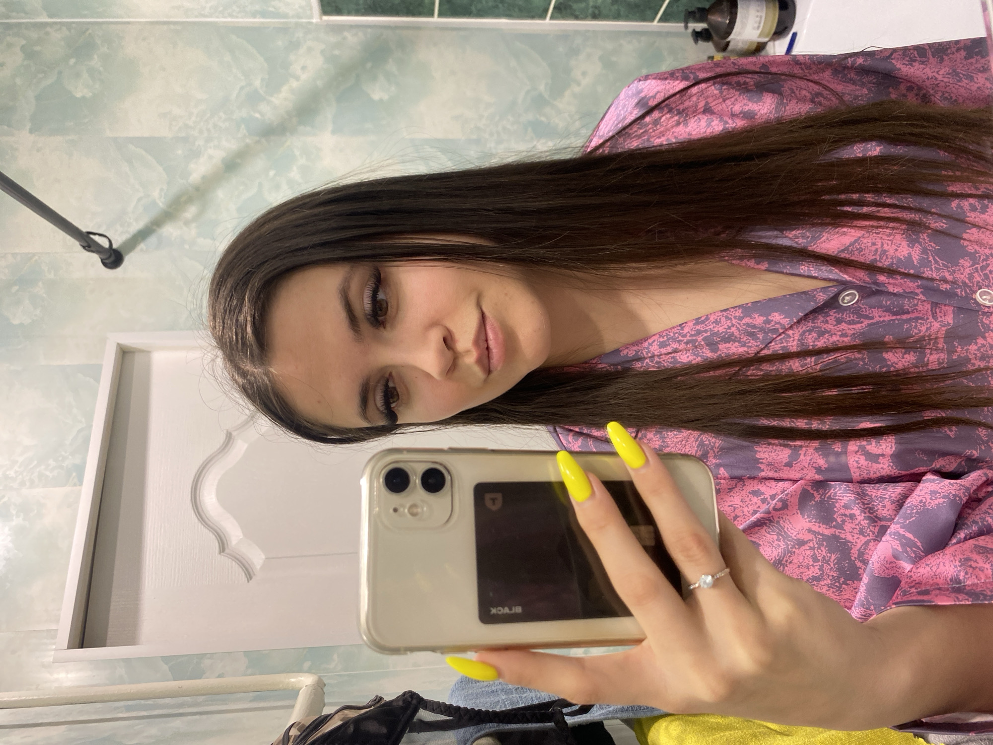 A woman is taking a selfie indoors. She is wearing a pink and purple patterned pajama top and has long brown hair. Her fingernails are painted bright yellow. She is holding a silver iPhone with a black card in the case. The background appears to be a bathroom with light green and white marble patterned tiles. A white door with decorative molding is visible. The location might be Chaikovsky, Russia.