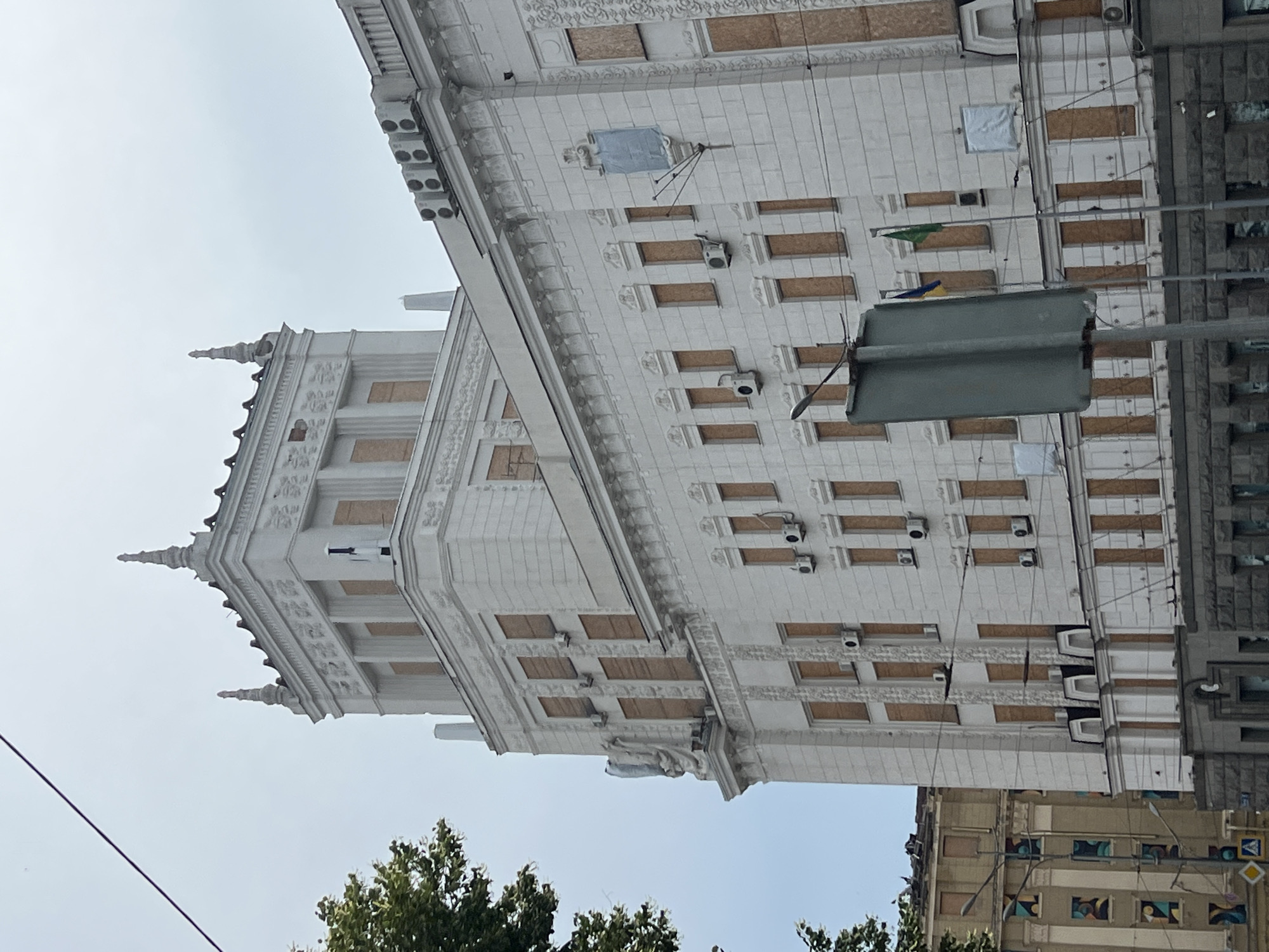 The image captures a side view of a large, ornate white building in Kharkiv, Ukraine. The building's windows are boarded up with plywood, suggesting damage or protection. Air conditioning units are visible mounted on the exterior walls. A prominent tower with spires rises from the top of the building. Decorative architectural elements, including ornate carvings and statues, adorn the facade. In the foreground, a street sign is visible. Two flags, one appearing to be the Ukrainian flag and another green flag, are mounted on poles in front of the building. To the left of the main building, another building with columns and colorful artwork on its windows is partially visible. The sky is overcast.