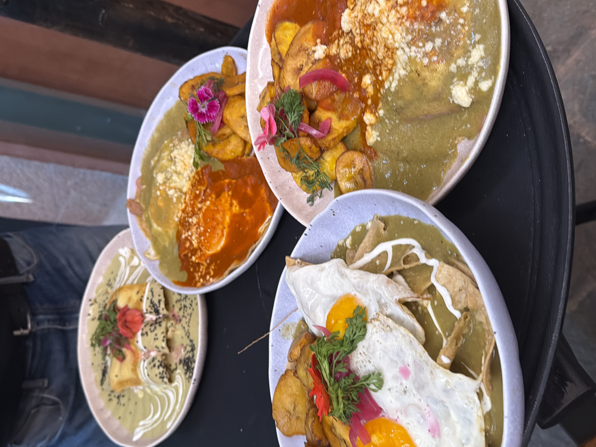 Here is a description of the image:
The image shows four plates of food, likely a variety of chilaquiles dishes. Each plate features a different combination of elements. All plates appear to be round with light-colored rims. The plates are positioned on a dark surface, possibly a table. The plates are arranged in a clustered formation.
The individual plates contain various components, including fried plantains, eggs (some appear fried with yolks intact), tortilla chips, and sauces in different colors, notably green and red. Some plates include crema or other white drizzles. Garnishes include a sprinkle of cheese and vibrant, small flowers and herbs.
One plate has a prominent stack of what appears to be fried cheese or tofu. Other plates have chilaquiles, with tortillas smothered in different sauces. A section of jeans is visible in the lower left portion of the image.
The location of this image is Unknown, Unknown.