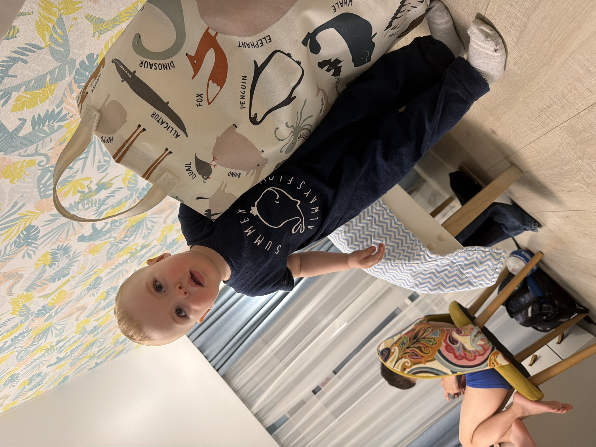 A toddler with blonde hair and blue eyes looks towards the camera, seemingly held or standing next to a large, cream-colored tote bag. The child is wearing a dark blue short-sleeved shirt with a white whale graphic and the partially visible text "SUMMER" and "ALWAYS FLO" (likely "ALWAYS FLOW" or "ALWAYS FLOATING"). Light grey socks cover their feet.
The tote bag, prominently featured, is adorned with various animal illustrations and their names in black text. Visible animals and text include "DINOSAUR", "ALLIGATOR", "FOX", "PENGUIN", "ELEPHANT", "WHALE", "RHINO", "QUAIL", "HIPPO", "MOUSE", "OCTOPUS", and partially visible "AMPHIBIAN" and "ANTELOPE".
The setting appears to be indoors, possibly a child's room or play area in Votkinsk, Russia. The background features a patterned wallpaper with light blue, yellow, and pink tropical leaf motifs. A light wooden floor is visible beneath the child. In the background, sheer white curtains hang over a window, and a wooden high chair or stool with a colorful, patterned cushion can be seen. Part of another person's legs, wearing blue shorts, is visible near the high chair, suggesting they might be sitting or kneeling. The overall lighting is bright, indicative of daytime. The child's expression is one of curiosity and engagement.