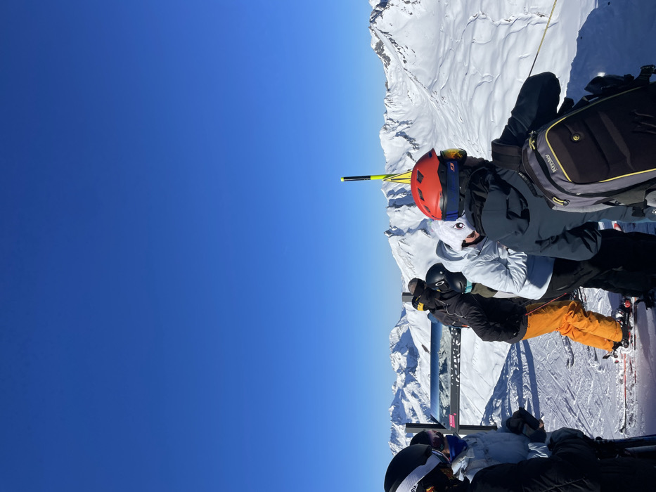 The image captures a vibrant winter sports scene high in the snow-covered mountains of Séez, France, under a brilliant, cloudless blue sky. The scene is bathed in bright sunlight, indicating excellent weather conditions for skiing or snowboarding.
In the mid-ground, a group of skiers and snowboarders is gathered on a snowy slope or platform, looking towards the expansive mountain range. They are all dressed in full winter sports attire, including helmets, goggles, and insulated jackets and pants.
Prominently featured is a person on the left wearing a bright red helmet with goggles, a grey ski jacket, and a black backpack displaying the "K2" logo. Adjacent to them is another individual in a white jacket and dark helmet. Further to the right, a person in a black jacket and distinctive bright orange ski pants stands on skis, which are partially visible and show the brand "NORDICA." Other individuals in dark ski gear are partially visible in the foreground, suggesting a larger group.
A yellow and black striped pole or marker stands upright in the snow behind the central group. A metal structure, possibly part of a railing, signpost, or terrain park feature, is also visible behind the people, with the word "SKATEBOARD" clearly printed on it. The background features endless snow-capped peaks and valleys, emphasizing the high-altitude, alpine environment. The clear visibility and bright conditions suggest a perfect day for mountain activities.