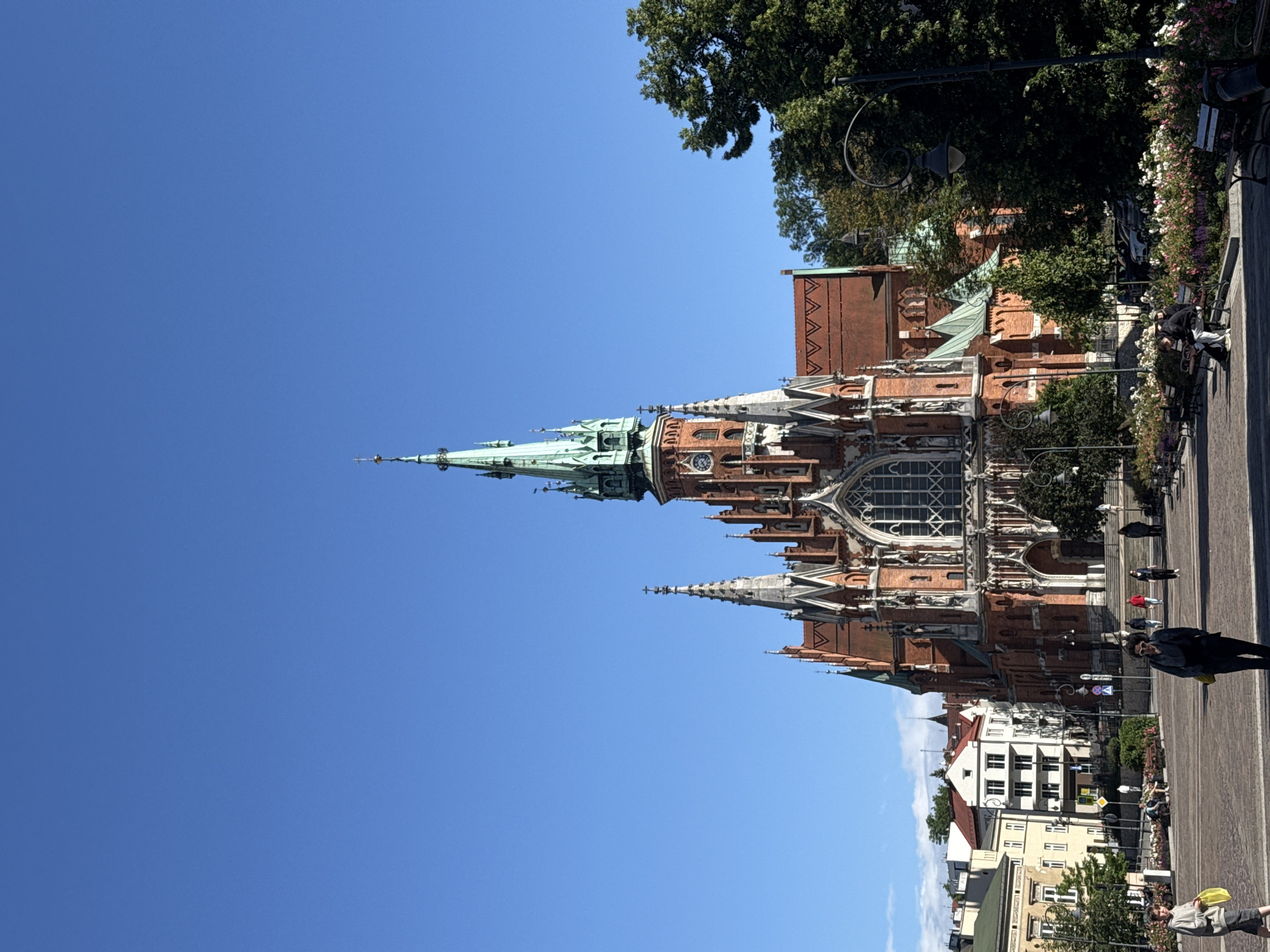 The image depicts a view of St. Mary's Basilica in Krakow, Poland, on a clear, sunny day with a bright blue sky. The basilica, a prominent Gothic church, is the central subject, characterized by its red brick facade, intricate spires, and a large rose window. The building's architecture features multiple towers, pinnacles, and decorative elements.
In the foreground, a cobblestone plaza is visible, with several people walking and sitting. To the left, a young person in a grey hooded sweatshirt and dark skirt holds a yellow bag. Further into the plaza, a person in a dark outfit with sunglasses and a yellow bag walks towards the camera. Other individuals are seen in the middle ground, some walking, others sitting on benches.
Flanking the plaza are buildings with traditional European architecture, including apartment buildings and what appears to be a smaller church or historical structure. Green trees, including a large, dense tree on the right side of the frame, provide a natural element to the urban landscape. Flowerbeds with various colorful flowers are also present along the edges of the plaza and near the benches. Streetlights with classic designs are placed along the pathways. The overall scene conveys a bustling yet serene atmosphere in a historic city square.