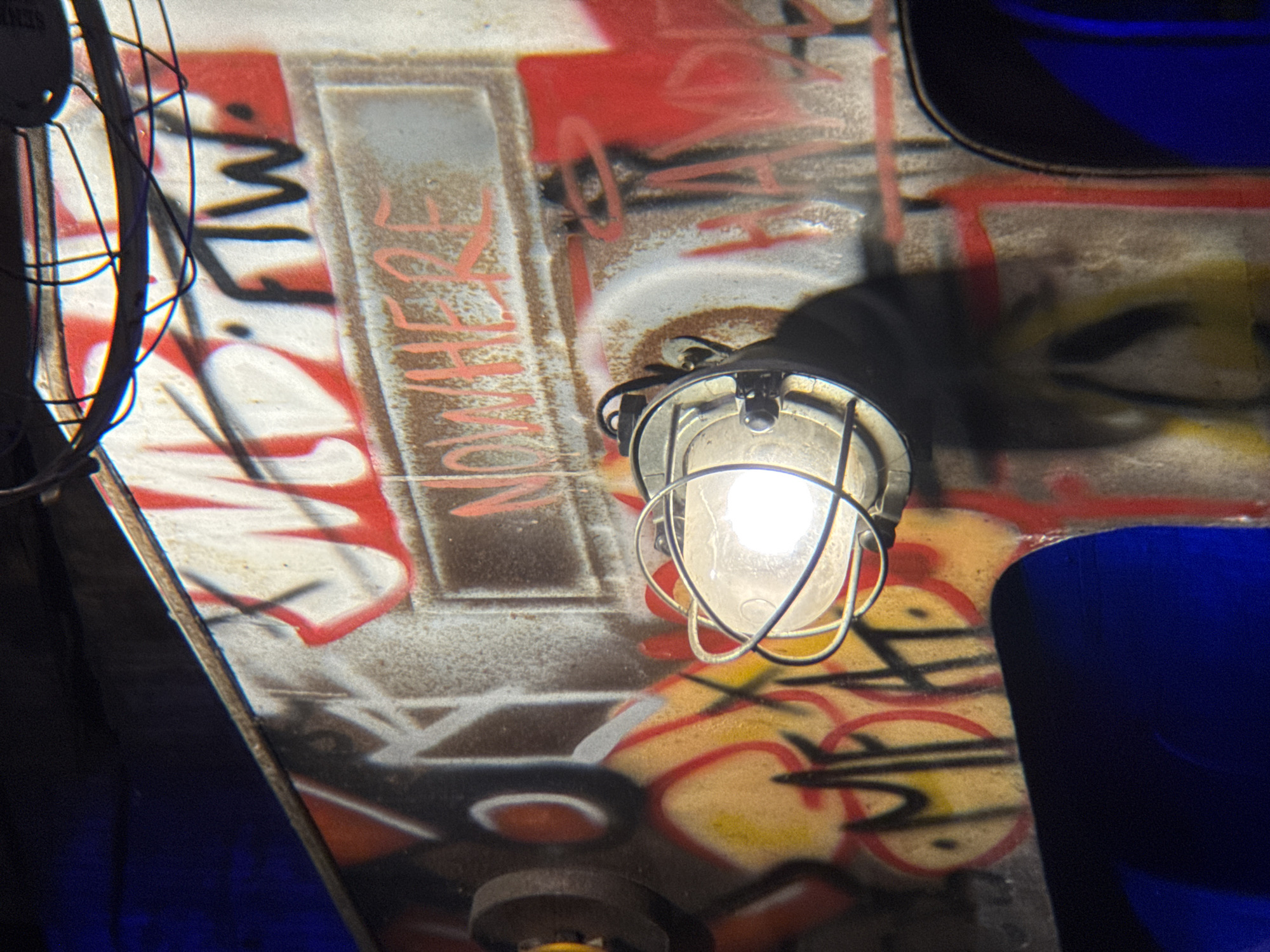 The image depicts a close-up, low-angle view of a weathered, graffiti-covered metallic surface, possibly part of an industrial structure or vehicle. A bright, bare light bulb encased in a wire cage illuminates the scene, casting a stark white glow and shadows. The graffiti is layered, featuring bold red and yellow spray paint alongside black lettering. Prominent text reads "NOWHERE" in red, and "FTW" in black. Other illegible graffiti is visible across the surface. The background is dark with an area of intense blue light in the lower left and right corners. The metallic surface shows signs of rust and wear. The location is identified as Thuận Hóa Ward, Vietnam. There are no people or animals visible in the image.