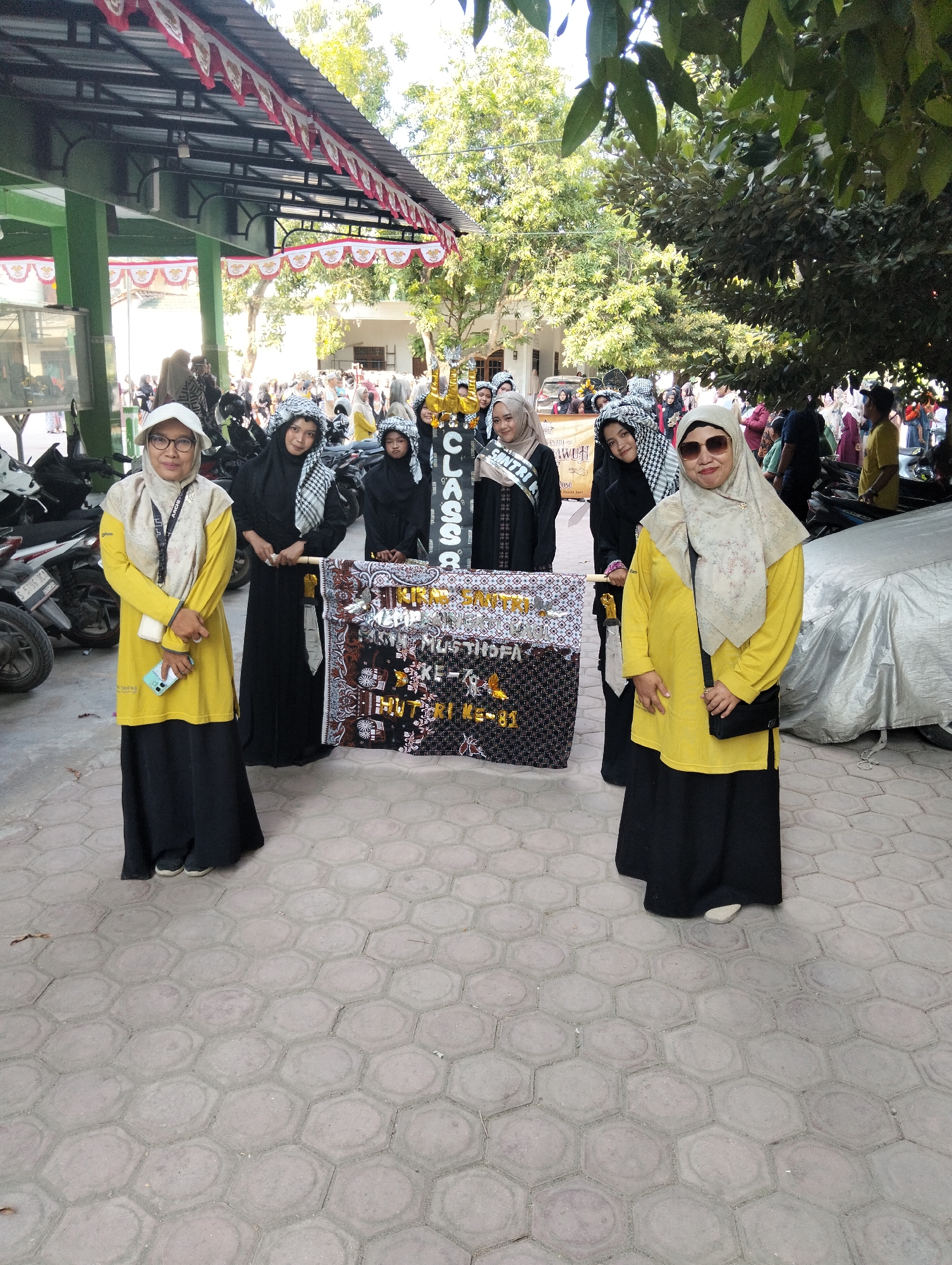 Six individuals, five women and one person of indeterminate gender, stand in front of a banner. The women are dressed in traditional Islamic attire, including hijabs and long dresses, with three of them wearing black attire and two in yellow and black outfits. The banner, held by two individuals on either side and supported by poles, displays text and decorative patterns. The scene appears to be outdoors, with a paved ground surface composed of hexagonal tiles. In the background, a building with a covered walkway and trees are visible, suggesting an urban or semi-urban environment. Several motorcycles are parked to the left, and a covered vehicle is parked to the right, partially obscuring the background. The overall lighting suggests it is daytime. The location is identified as Kranji, Indonesia.