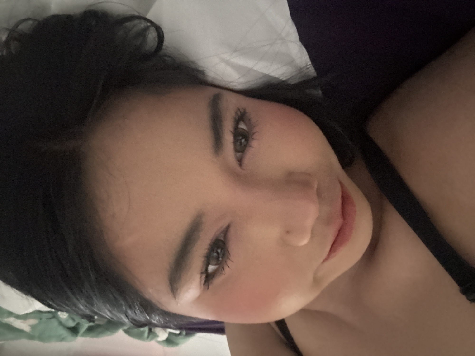 In North Jakarta, Indonesia, the image captures a close-up, slightly tilted view of a young adult female lying down and looking directly into the camera. The subject has dark hair, dark shaped eyebrows, visible mascara on long eyelashes, and brown or hazel eyes. Her lips are slightly parted, revealing a pinkish-red tint. Pink blush is visible on her cheeks. A dark (likely black) strap, consistent with a bra or top, is prominent along the left side of the frame. The subject is resting on white bedding, with a dark purple or blue fabric visible on the lower left. The background is shallowly focused, showing a blurred green textile object in the upper right. The overall lighting is soft and even.