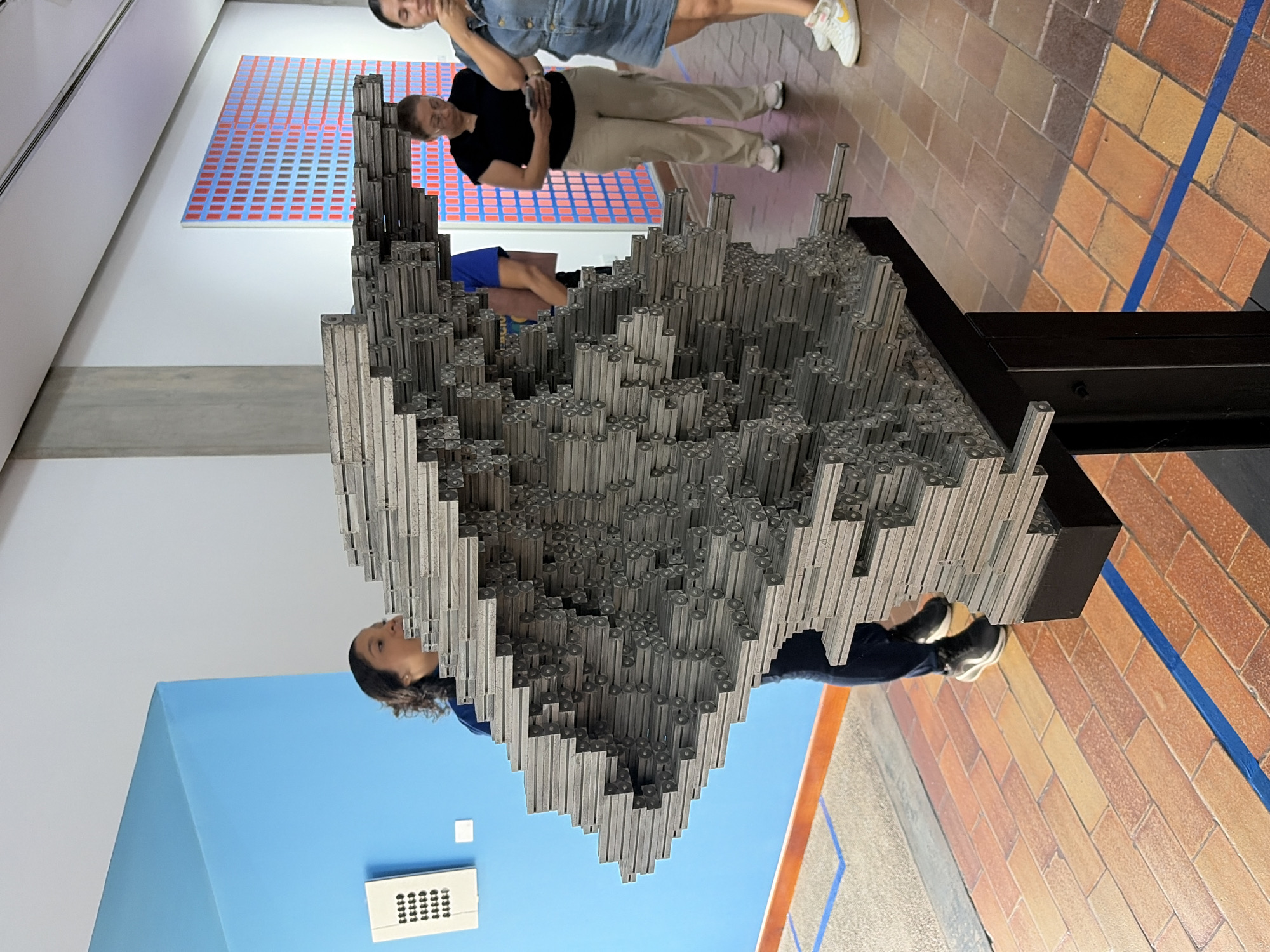 The image shows an art installation, likely an exhibit in a gallery or museum. The installation is a three-dimensional sculpture composed of many small, gray, square-shaped bars or tubes, stacked together to form a complex, multi-tiered structure. The sculpture sits on a black, rectangular base.
Several people are in the vicinity of the art piece. One woman is looking at the piece. Another is standing further away, looking at the sculpture. Behind these women, a framed artwork with a grid pattern of red and blue squares is hung on the wall.
The environment consists of an art gallery or museum setting. There is a blue wall in the background, with an air vent visible. The floor is made of orange-brown brick tiles. The image provides insufficient information to determine whether this is Ciudad Bolivar, Venezuela.
