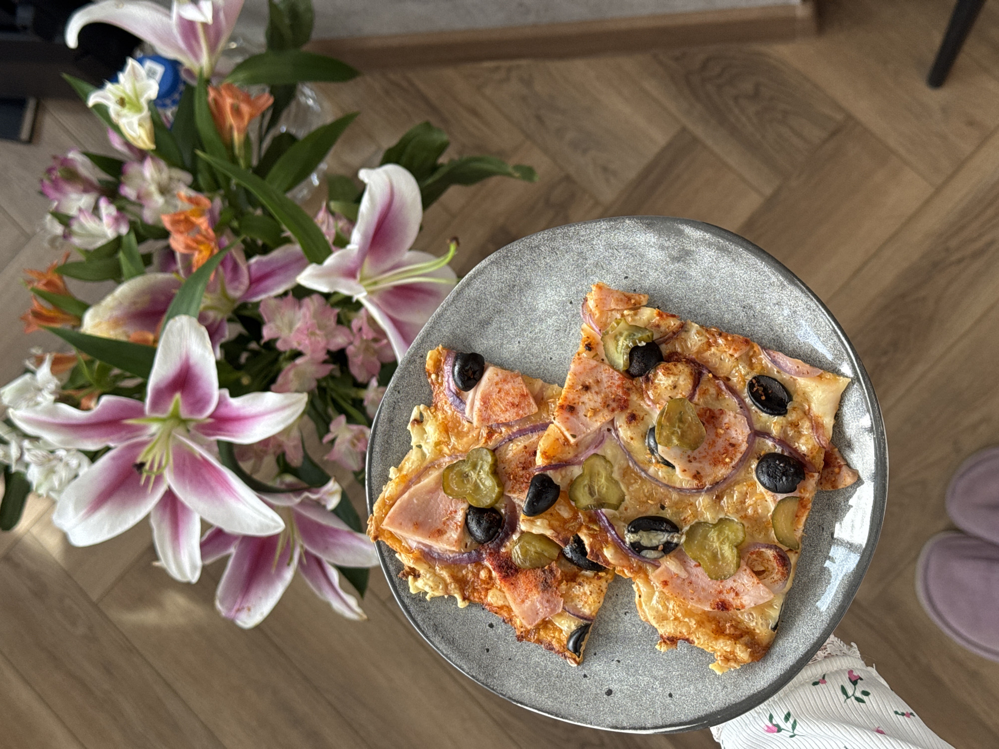The image captures a top-down view of a culinary still life, potentially indoors. A plate containing two pizza slices dominates the central space. The pizza features toppings like ham, red onion rings, pickles, and black olives. The plate is a dark gray with a speckled texture.
Adjacent to the plate, towards the left, is a vibrant bouquet of flowers, including lilies with white and pink petals, alongside other floral arrangements in shades of white, pink, and orange.
The background features a light brown, wood-patterned surface, presumably a tabletop. A partial view of a person wearing floral patterned sleeves and purple shoes appears in the lower right corner.
The scene appears to be situated in an unknown location, possibly a private residence in Unknown.