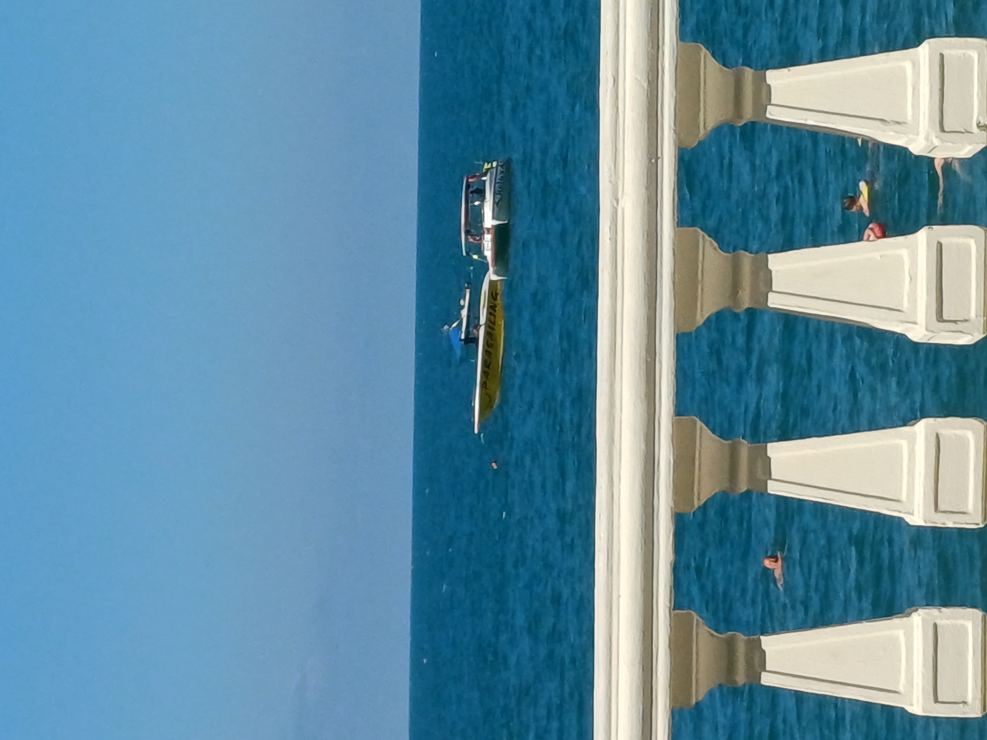 The image captures a bright, sunny day at sea, likely in Sochi, Russia, given the context. The scene features the azure blue water extending to the horizon, with a clear, light blue sky above. In the foreground, a white balustrade with decorative columns partially obscures the view of the sea.
On the water, two boats are visible. The first is a white boat with a red stripe, carrying several people. The second boat is long and yellow, appearing to be a type of raft or inflatable boat, with people on it. Further out in the water, two individuals are seen swimming, one with a yellow flotation device and the other in a red life vest.
The overall impression is one of a recreational activity on a warm day, with people enjoying the ocean. The sunlight suggests it is daytime, likely mid-morning to afternoon. There is no discernible text visible in the image. The presence of swimmers and boats points to a coastal or resort area.