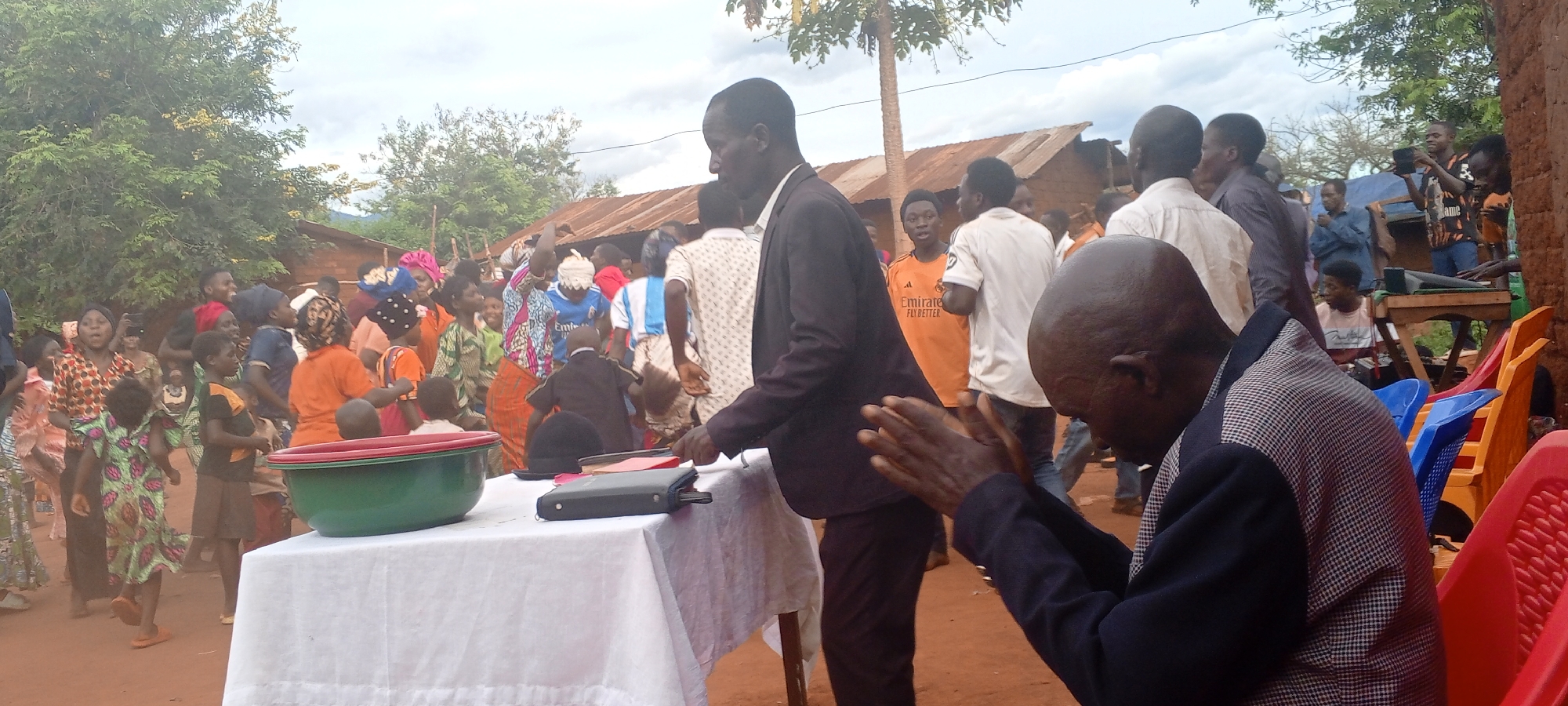 The image shows a gathering of people in Ngalula, Democratic Republic of the Congo. A crowd of adults and children stands and moves in the background. Some are dressed in colorful patterned clothes. A table with a white tablecloth is set up in the foreground. A green and red plastic basin, a book, and a black object are on the table. A man in a dark suit appears to be walking towards the table. In the lower right, a seated man with a shaved head is clapping his hands. Several plastic chairs, some red and some blue, are stacked behind him. A corrugated metal-roofed structure is visible in the background. The sky is overcast.