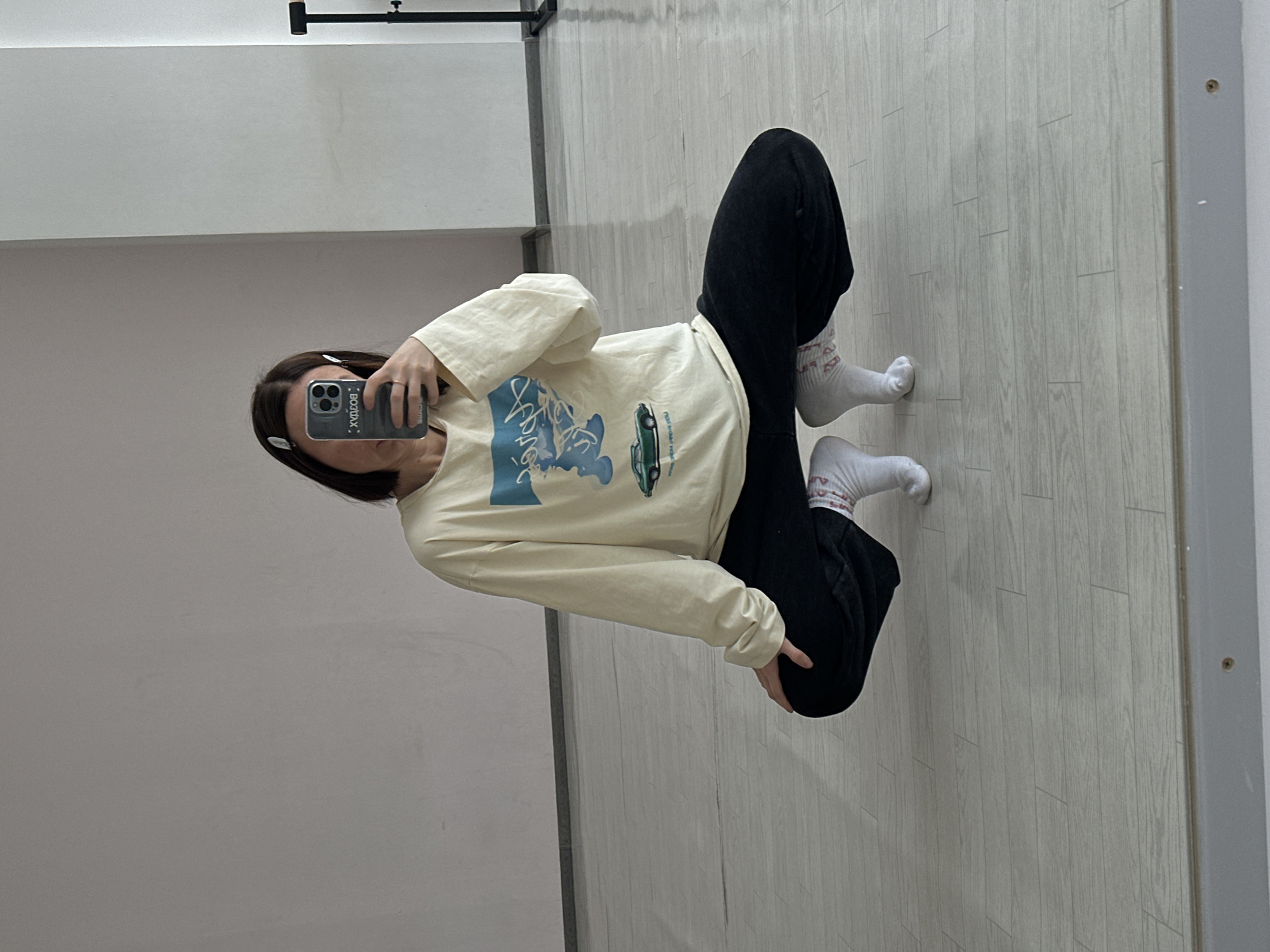 A woman is taking a selfie in a room. She is wearing a light-colored, long-sleeved shirt, dark pants, and white socks. She is holding a smartphone in her left hand. The room has light-colored walls and a light wood-patterned floor. A horizontal bar is visible on the wall. The location is unspecified.