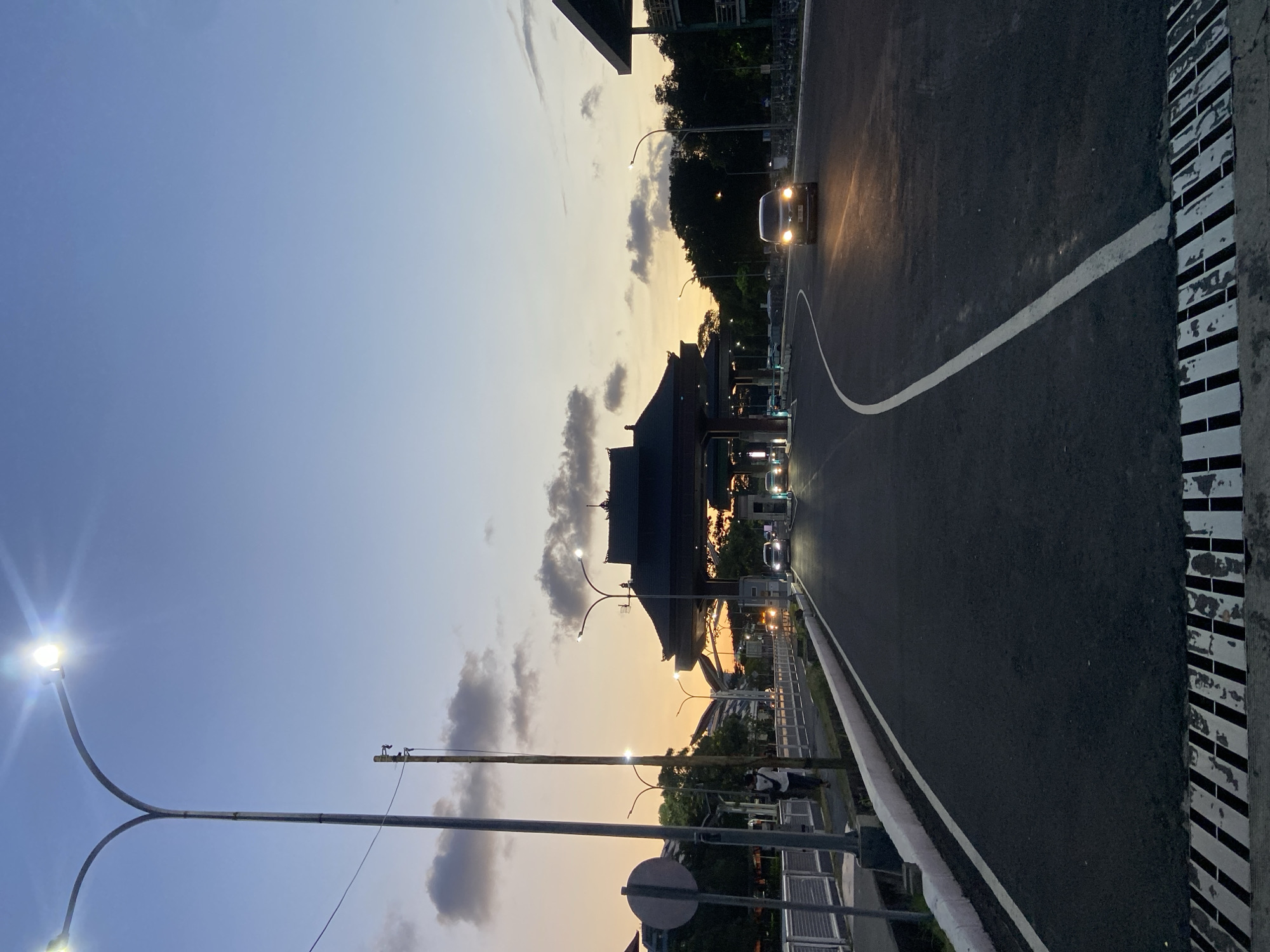 The image depicts a road at dusk in Kuta, Indonesia. A traditional Balinese gate-like structure stands in the middle distance, with trees and buildings on either side. Cars with headlights on are moving along the road. Streetlights illuminate the scene. The sky transitions from blue to orange.