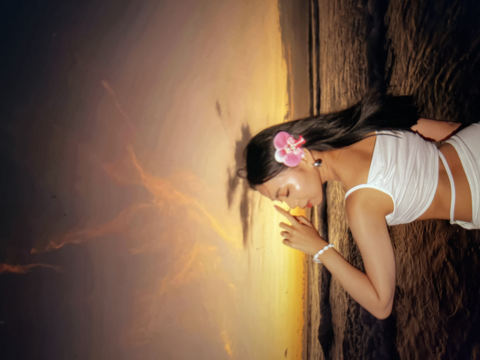 A young woman with long, dark hair adorned with a pink flower is standing on a beach at sunset. She is wearing a white top and bottom, and her right arm is raised with her hand near her face. The ocean is behind her, with waves crashing on the shore. The sky is a vibrant mix of orange, yellow, and purple, with wisps of clouds. The image appears to be taken in Cirahayu, Indonesia, given the beach setting and tropical flora.