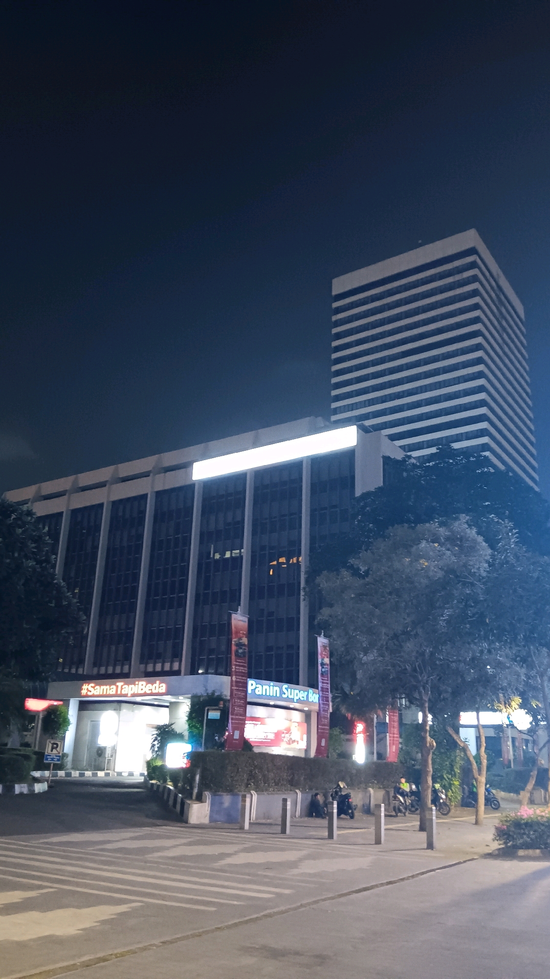 The image depicts the Panin Super Box building in Central Jakarta, Indonesia, at night. The building features a modernist architectural style with a prominent horizontal striped design on its upper sections. The lower facade of the building is illuminated, and signage is visible, including "#SamaTapiBeda" and "Panin Super Box." A parking sign with the letter "R" is present. Several trees with dense foliage are situated in front of the building. The foreground shows a street with visible lane markings, and several motorcycles are parked or stopped. A few individuals are seen near the motorcycles. The sky is dark, indicating nighttime.