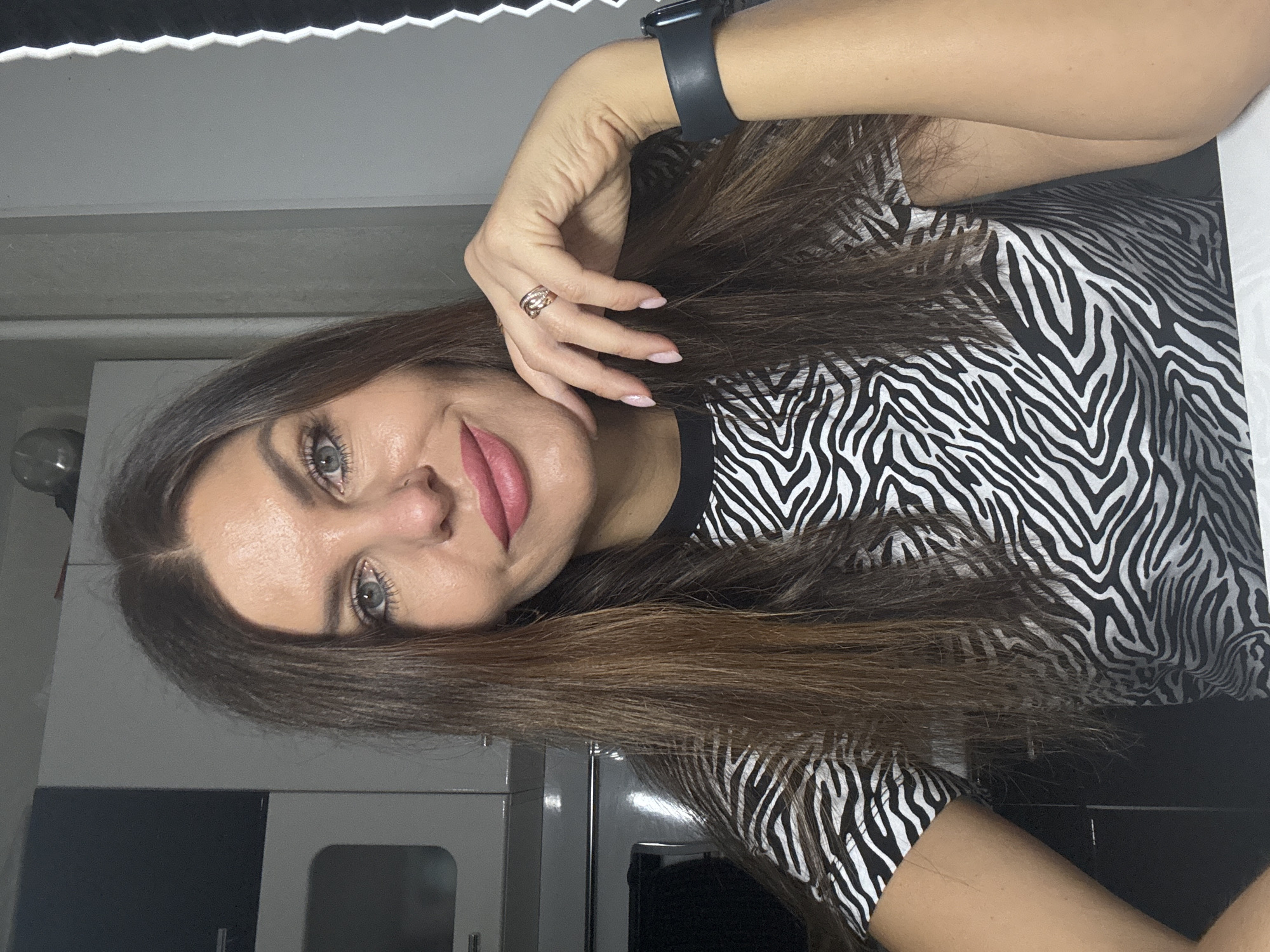 A woman is facing the camera. She has light gray eyes, brown hair, and wears pink lipstick. A ring is visible on her left ring finger and a smart watch on her left wrist. She is wearing a black and white zebra-print shirt. She appears to be inside a kitchen. Based on the prompt, the location is the city of Chaikovsky, Russia.