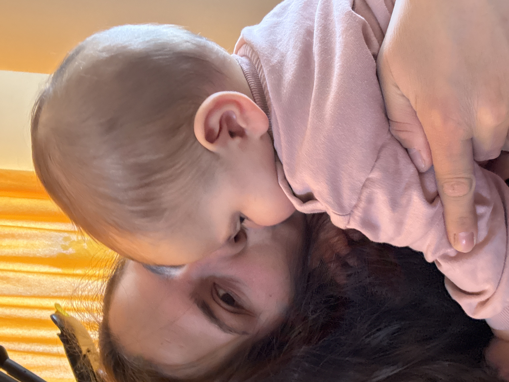 A close-up photograph shows an adult and a baby. The adult is lying down, facing upward, with dark, curly hair visible at the bottom of the frame. The baby, dressed in a pink long-sleeved shirt, is positioned on top of the adult, with the side of their face pressed against the adult's forehead. The baby's ear is visible. The adult's right eye is open, looking slightly to the right. A hand is visible near the baby's shirt. The background includes a yellow-toned curtain or blind. The location is not discernible from the image. No landmarks identifying the location as Sofia, Bulgaria are present.