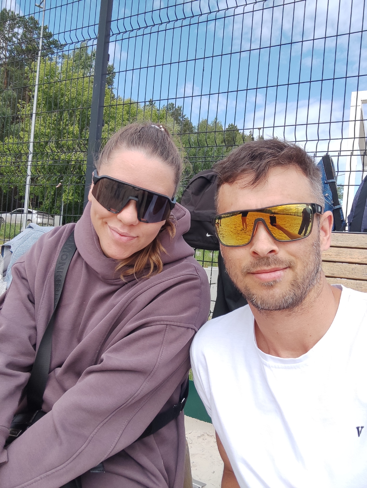 Two individuals are sitting on a wooden bench, posing for a selfie. The woman on the left is wearing a mauve-colored hoodie, and large dark sunglasses. A bag with a strap is visible across her torso. The man on the right is wearing a white t-shirt and sunglasses with mirrored lenses. Behind them is a fence, trees, and blue sky with clouds. Several bags and items are on the bench behind them. The location is outdoor in a park or recreational area.